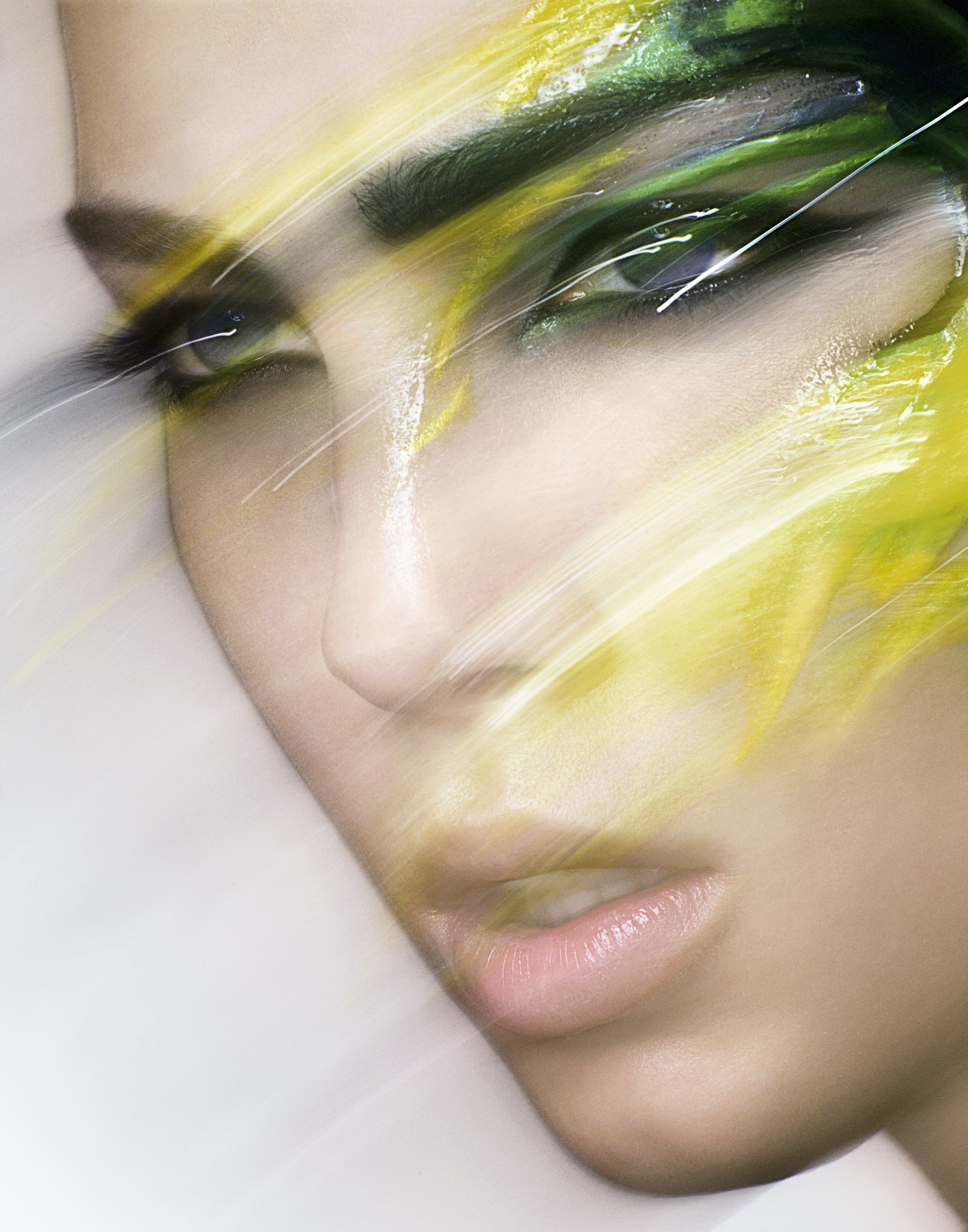 Close-up of a woman's face with vibrant green and yellow makeup, blurry streaks across the image adding an artistic effect.