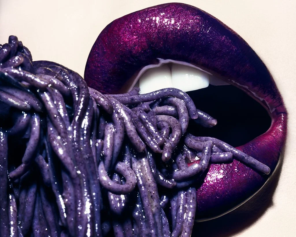 Close-up of lips with purple lipstick, with purple spaghetti noodles near the open mouth.