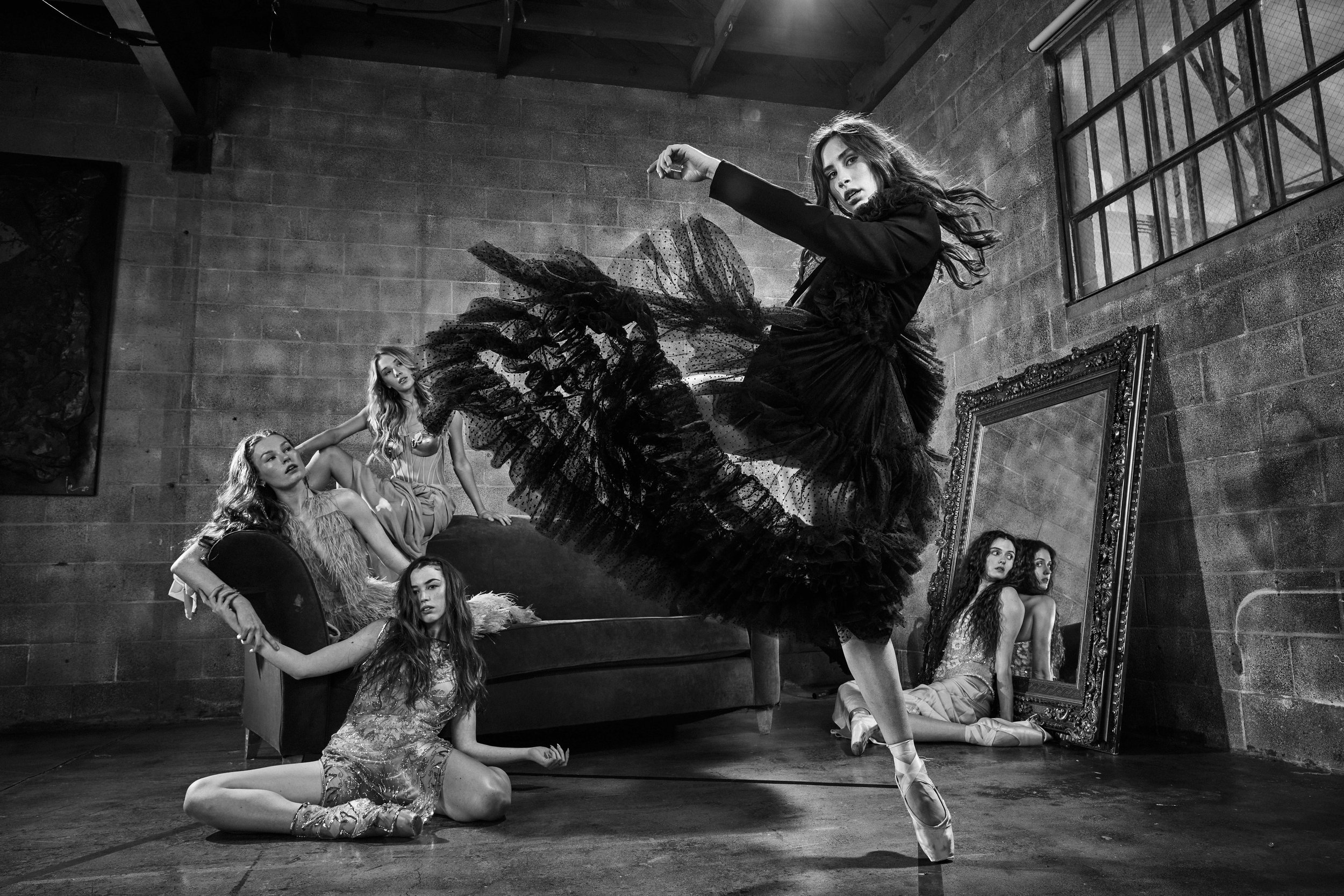 Black and white photo of five women in a studio with an industrial brick wall. One woman in the foreground, wearing a dark dress, is dancing en pointe. Three women sit or kneel on a couch, and one woman sits on the floor next to a large mirror on the