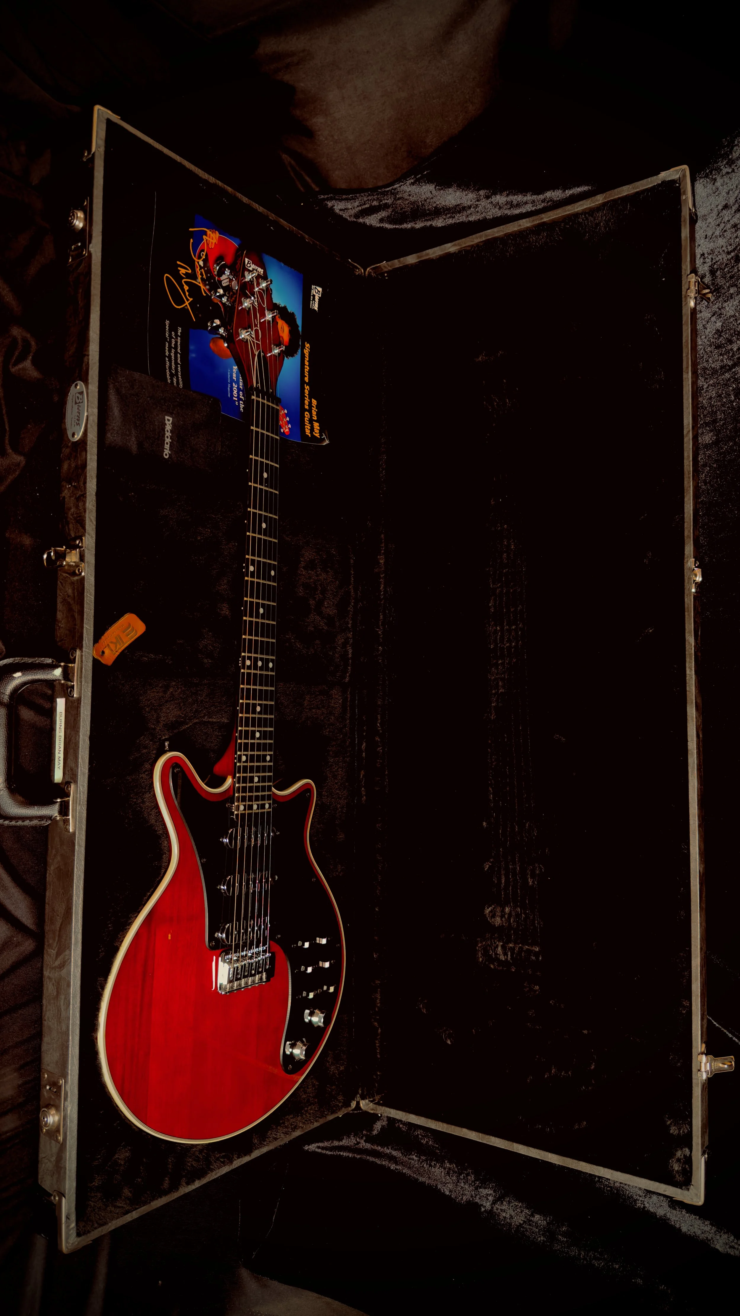 A red electric guitar with black pickguard and silver hardware sits in an open hard case lined with black velvet. There is a postcard of Eric Clapton and a tag inside the case.