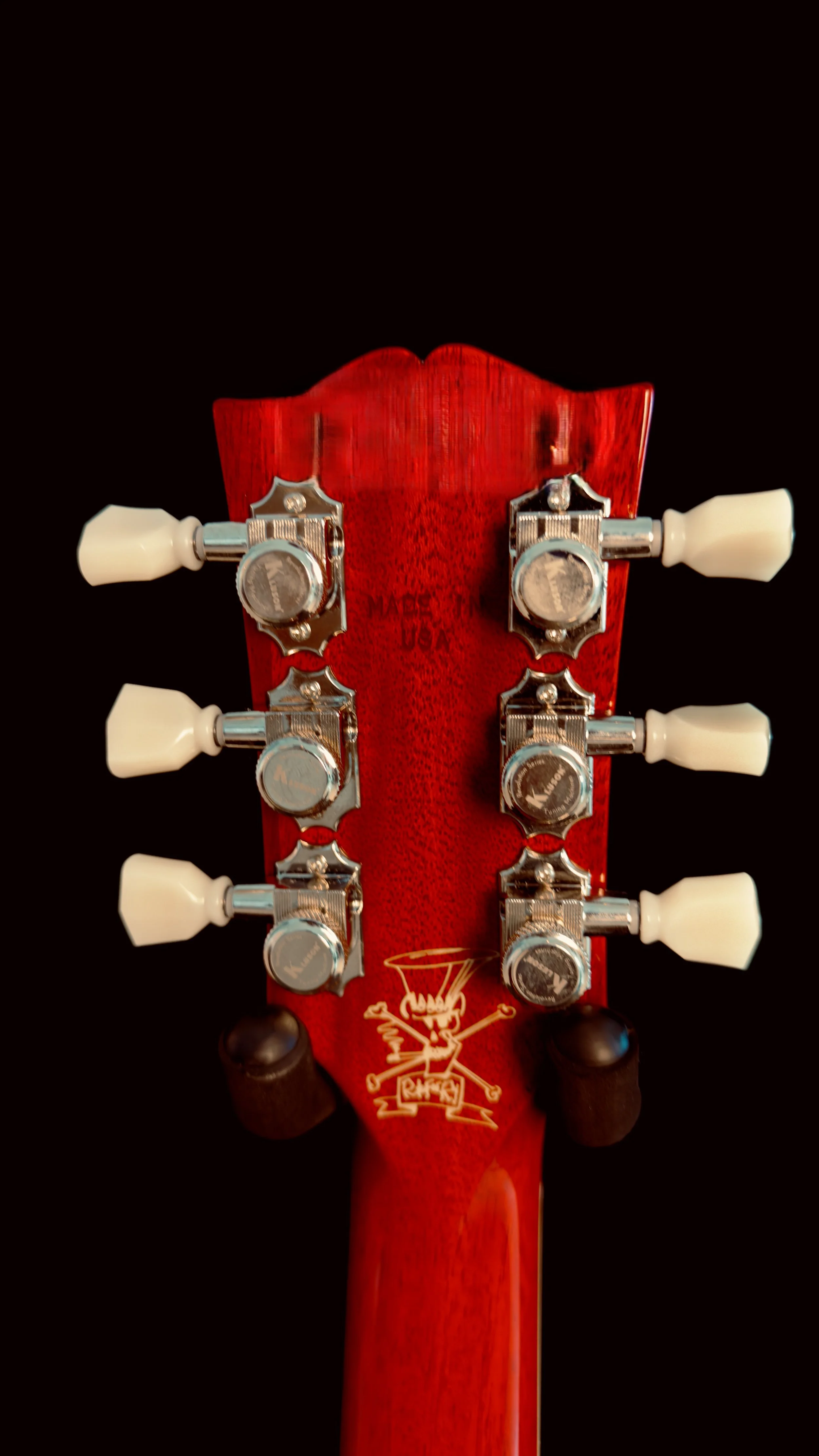 Close-up of the headstock of a red guitar with six tuning pegs, three on each side, and a small graphic of a devil with a pitchfork on it.