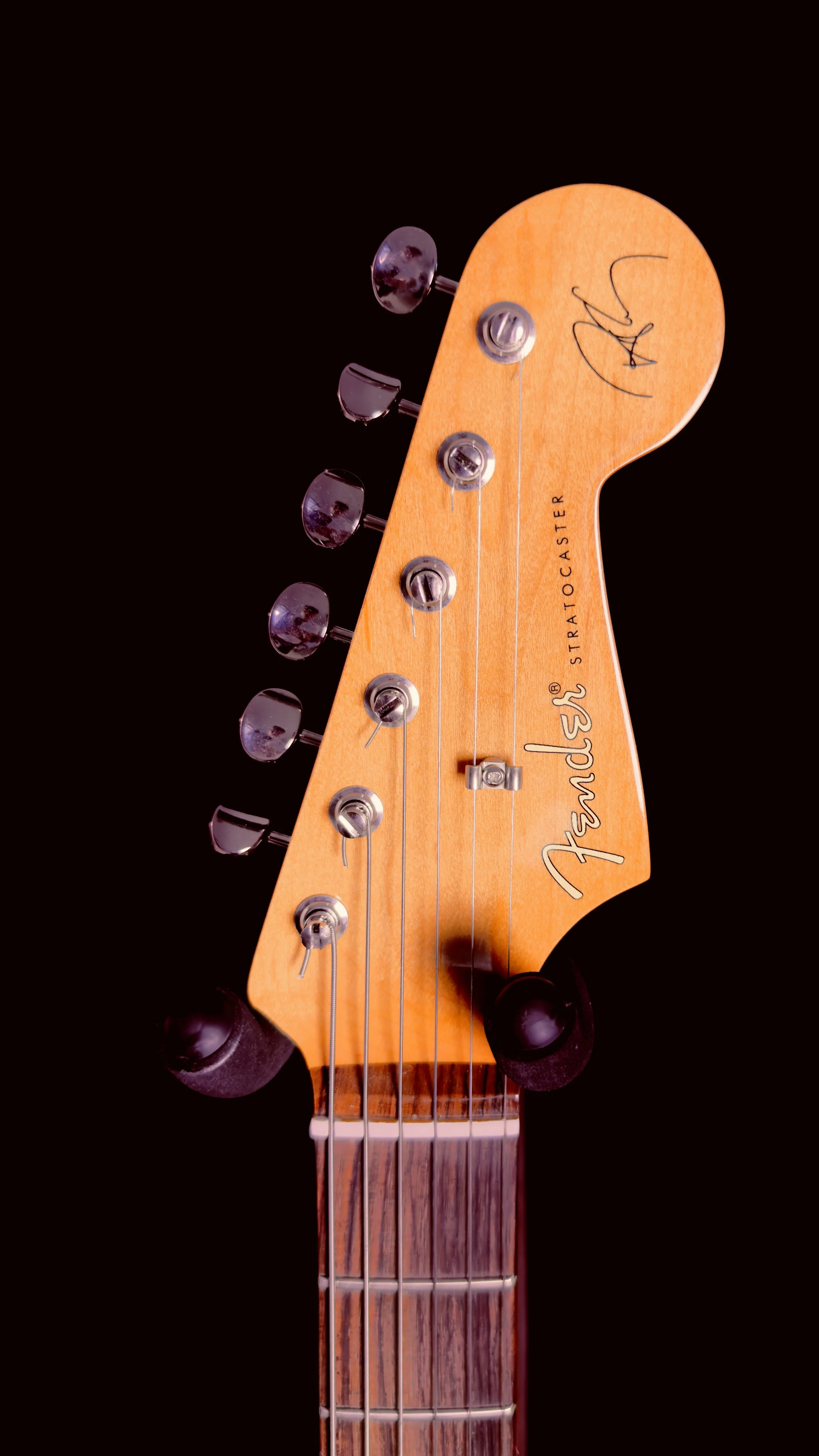 Close-up of a Fender Stratocaster guitar headstock with tuning pegs, strings, and a signature on the top right.
