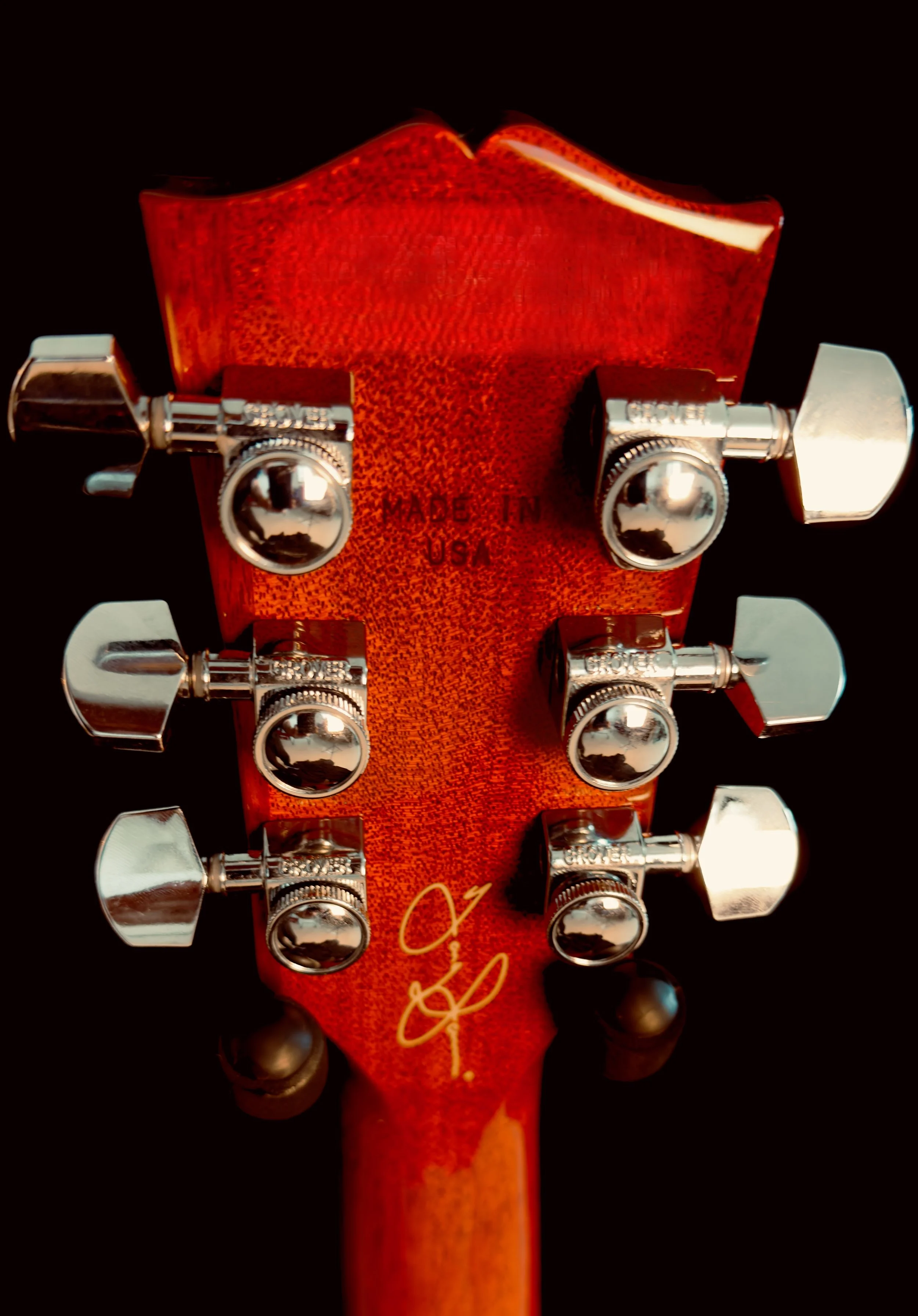 Close-up image of a red guitar headstock with six chrome tuning pegs and a signature near the bottom.