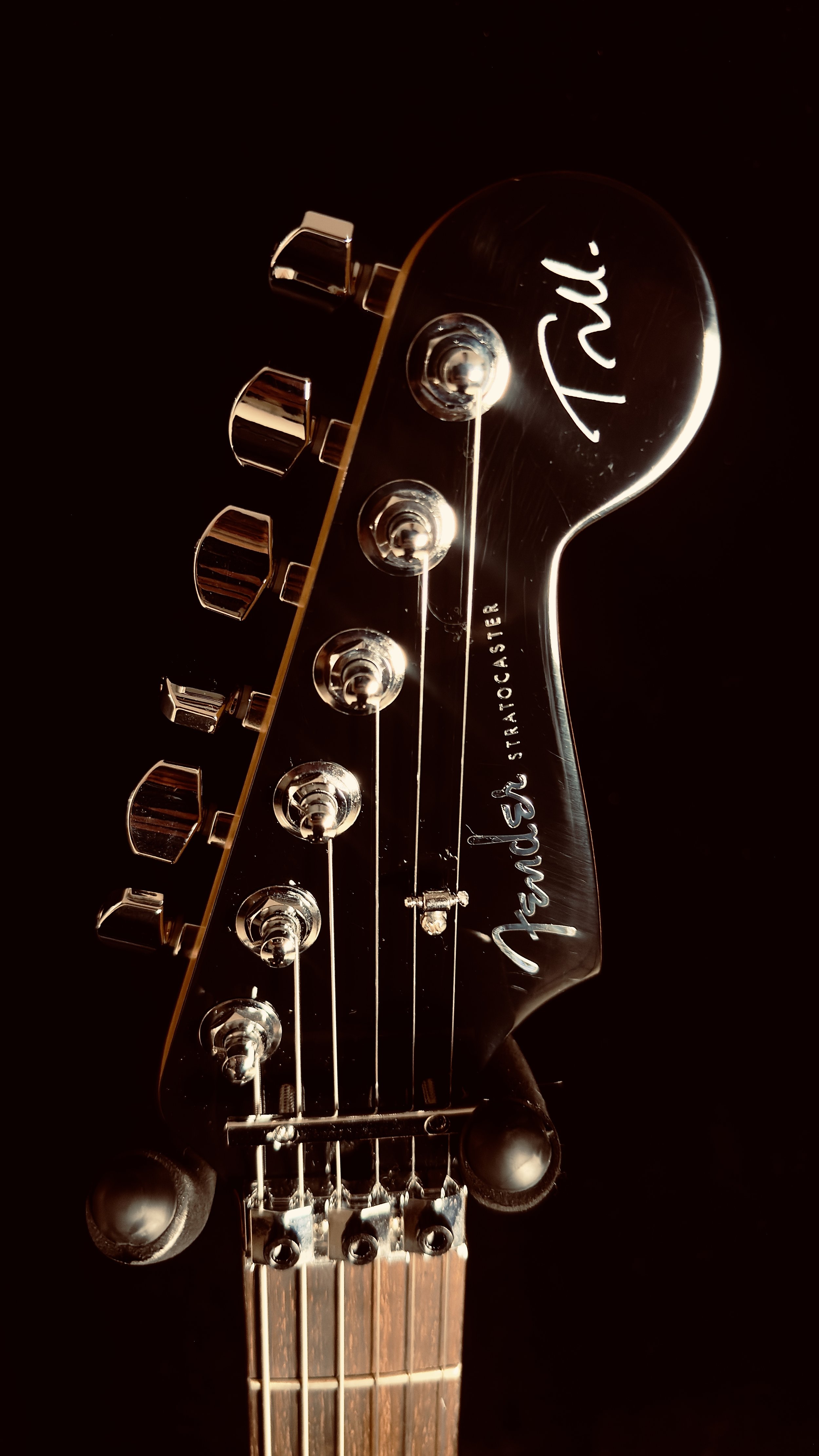 Close-up of the headstock of a Fender Stratocaster electric guitar, showing tuning pegs, strings, and the Fender logo.