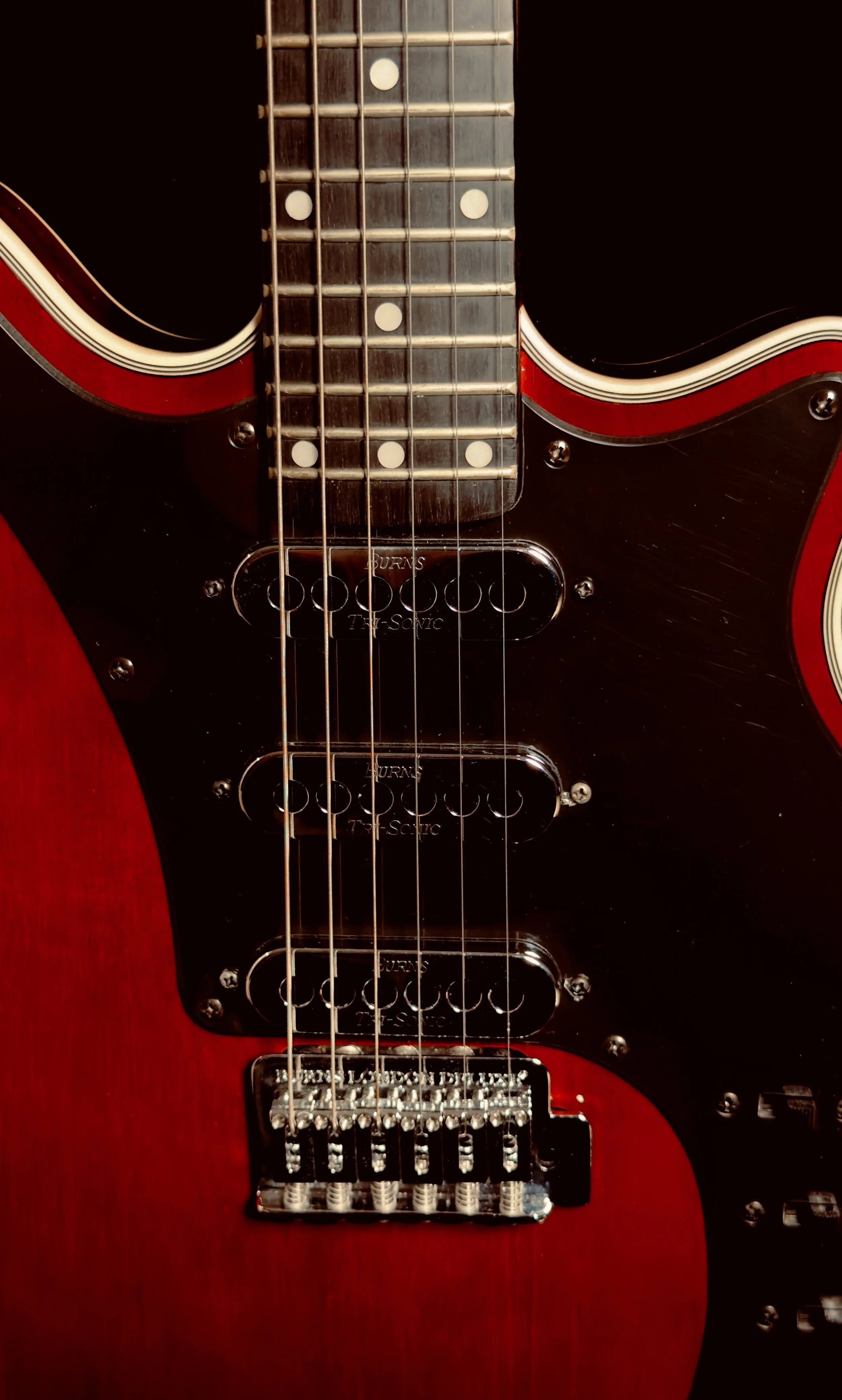 Close-up of a red electric guitar with a black pickguard, three pickups labeled 'Burns 7T-Servo', a whammy bar, and part of the fretboard with fret markers.