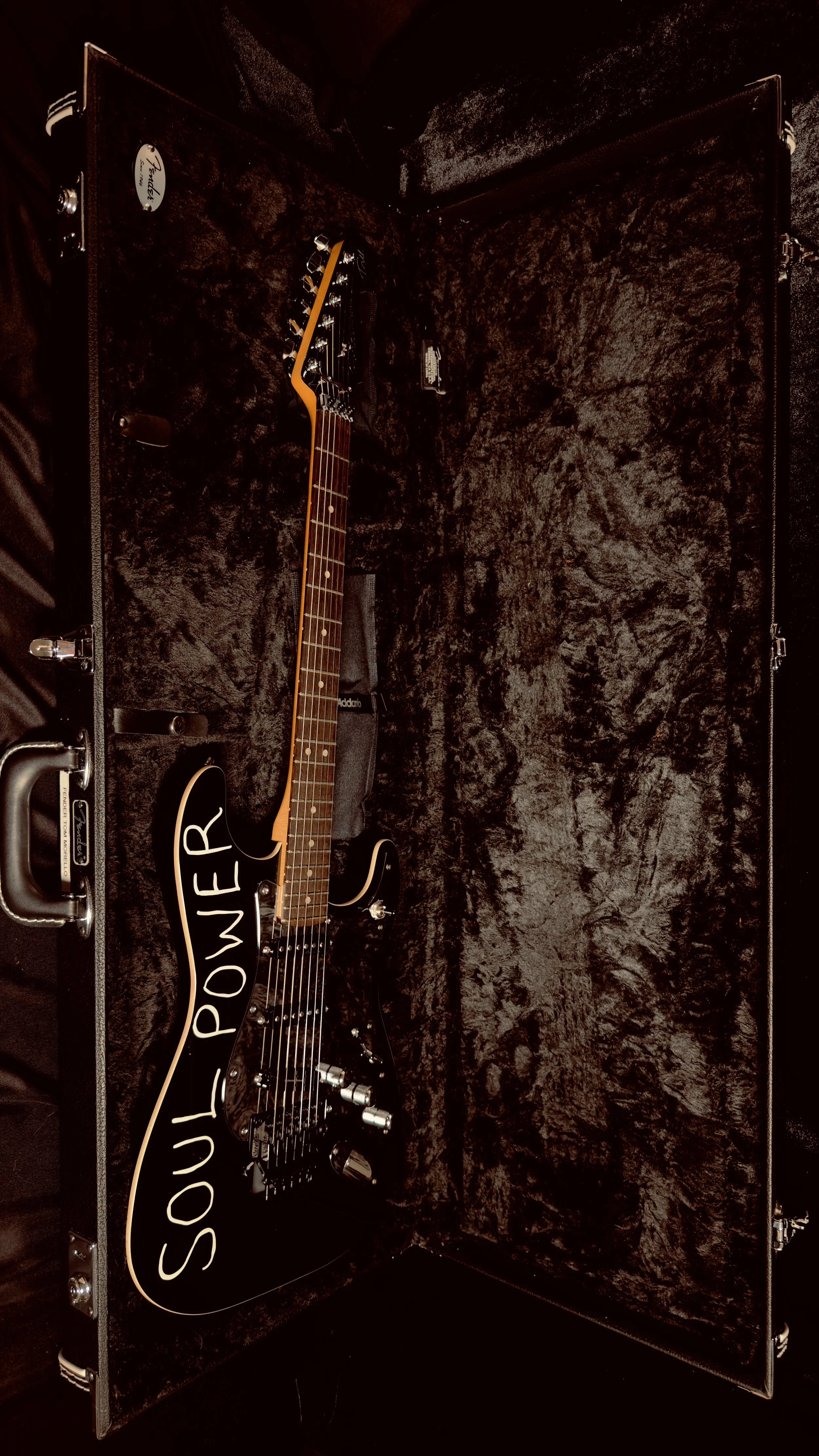 An electric guitar with a black body and a wooden neck inside a protective case lined with black plush. The guitar has a speech bubble shape with the words 'SOUL POWER' written on it in white. The case is open, revealing the guitar and some accessories.