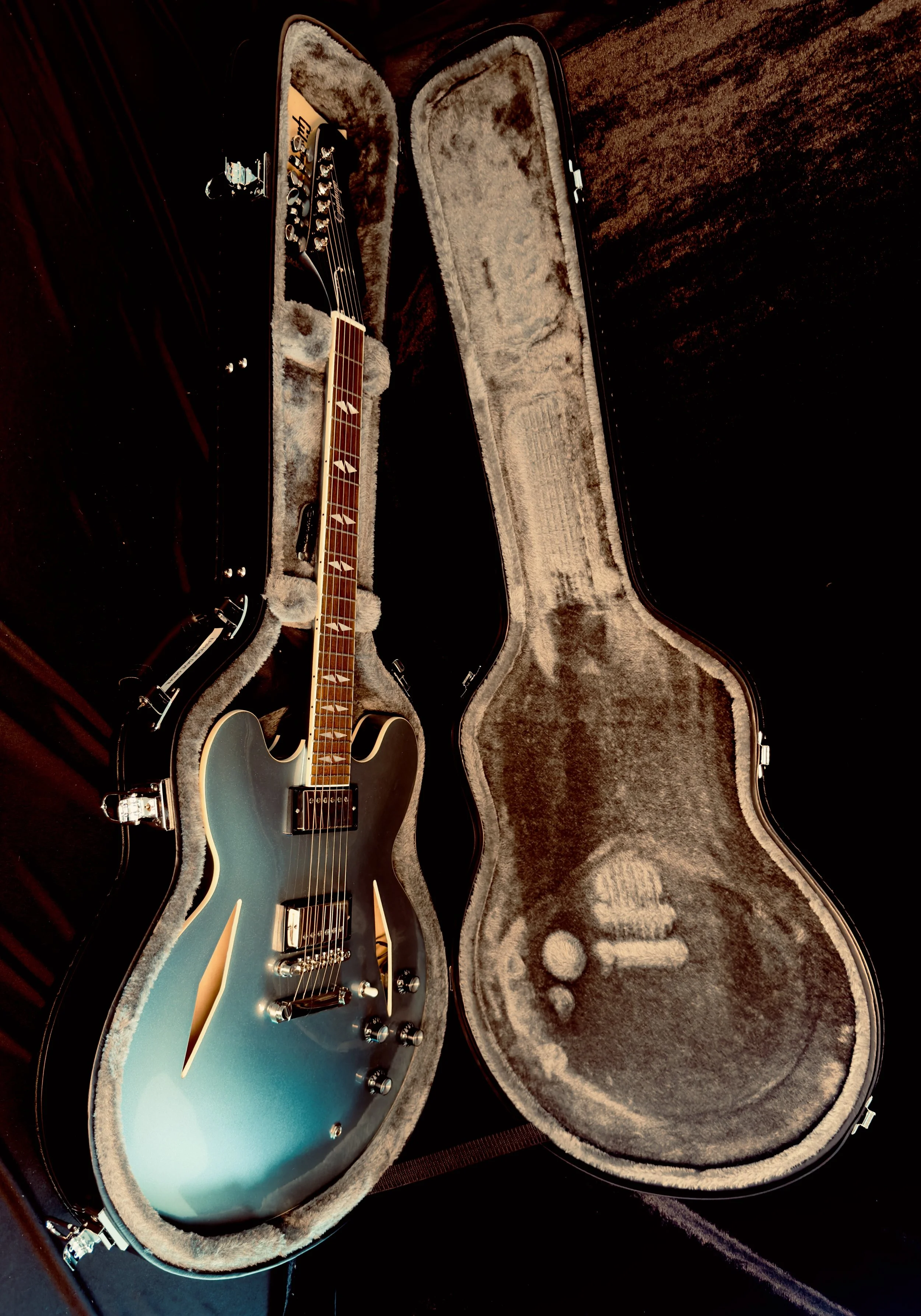 A blue electric guitar inside an open black hard shell guitar case with a plush interior, resting on a dark wooden surface.