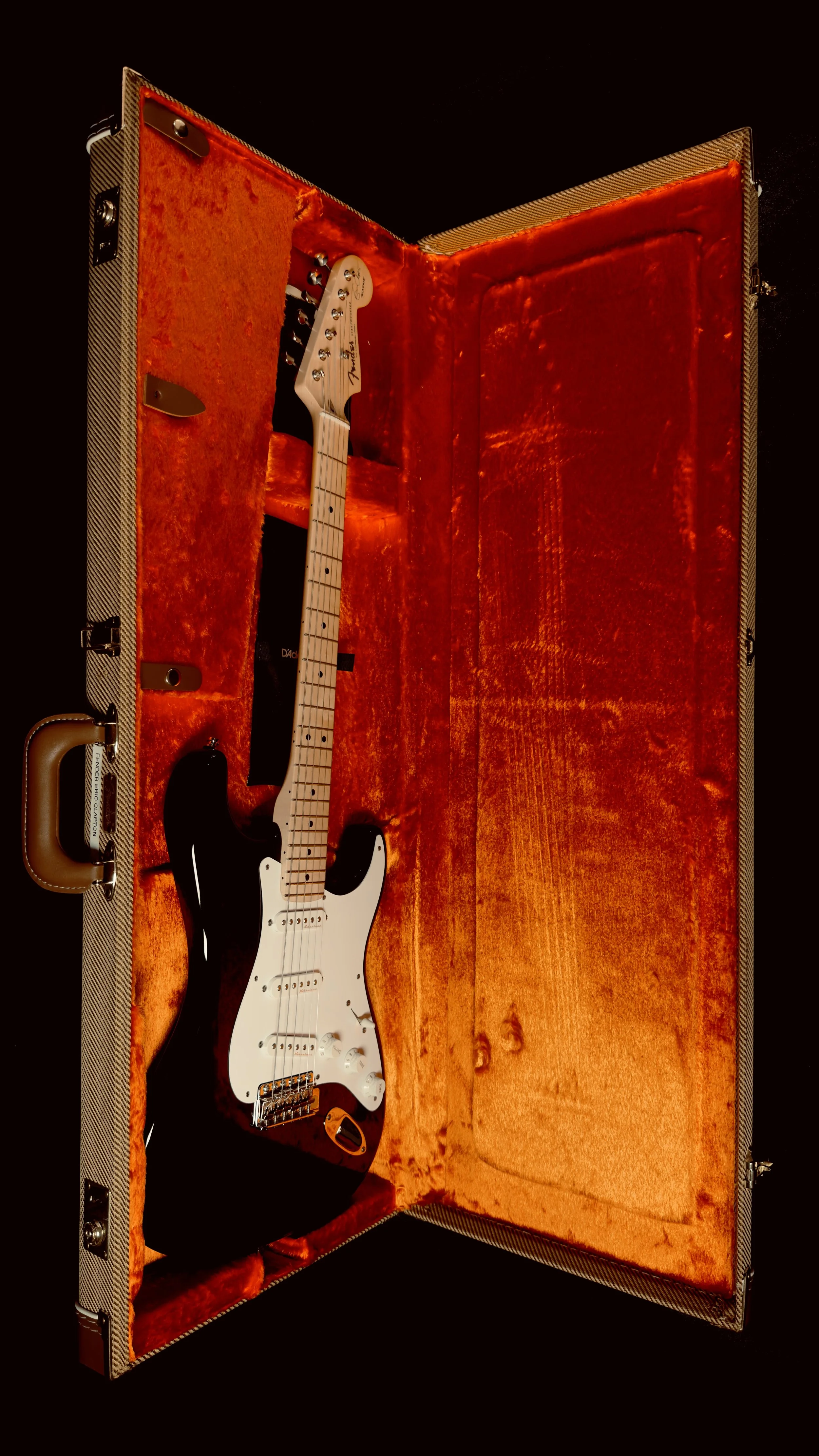 A Fender Stratocaster electric guitar with a black body and white pickguard inside an orange velvet-lined hard case.