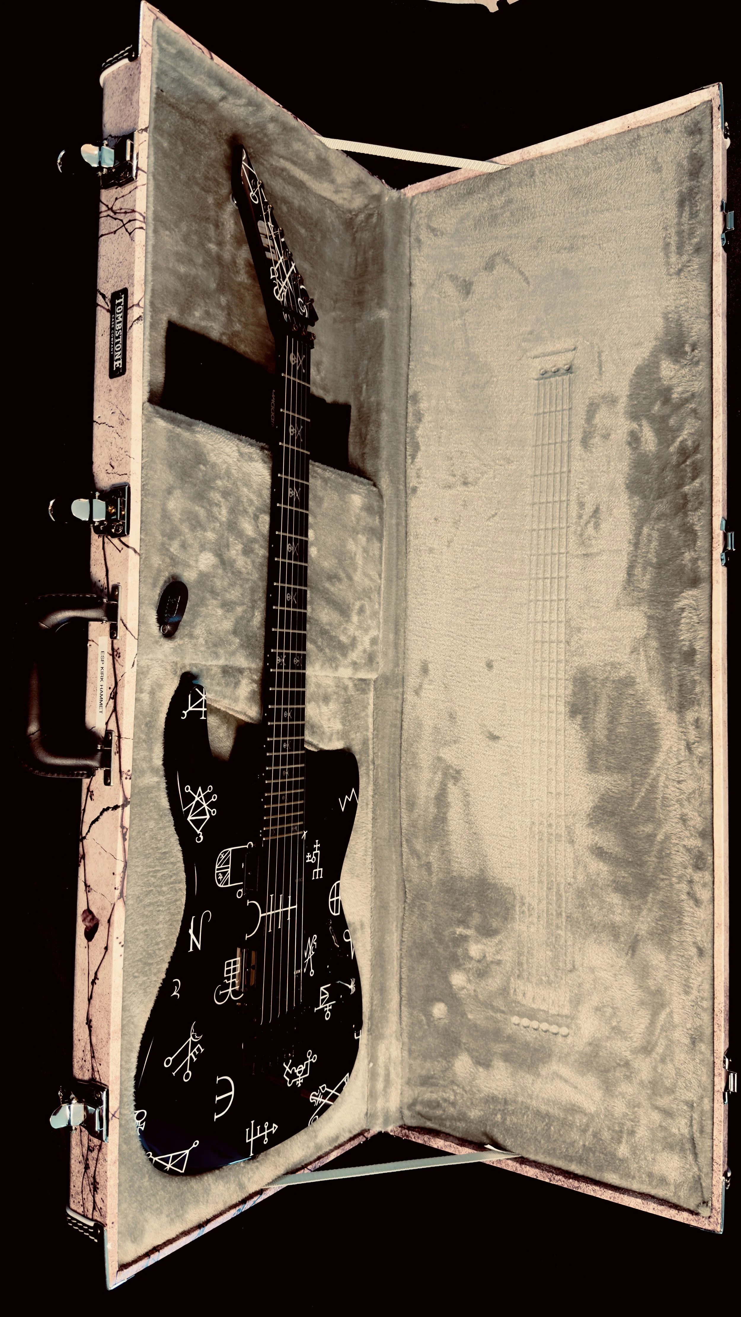A black electric guitar with white symbols and markings inside an open vintage pink guitar case with plush interior.