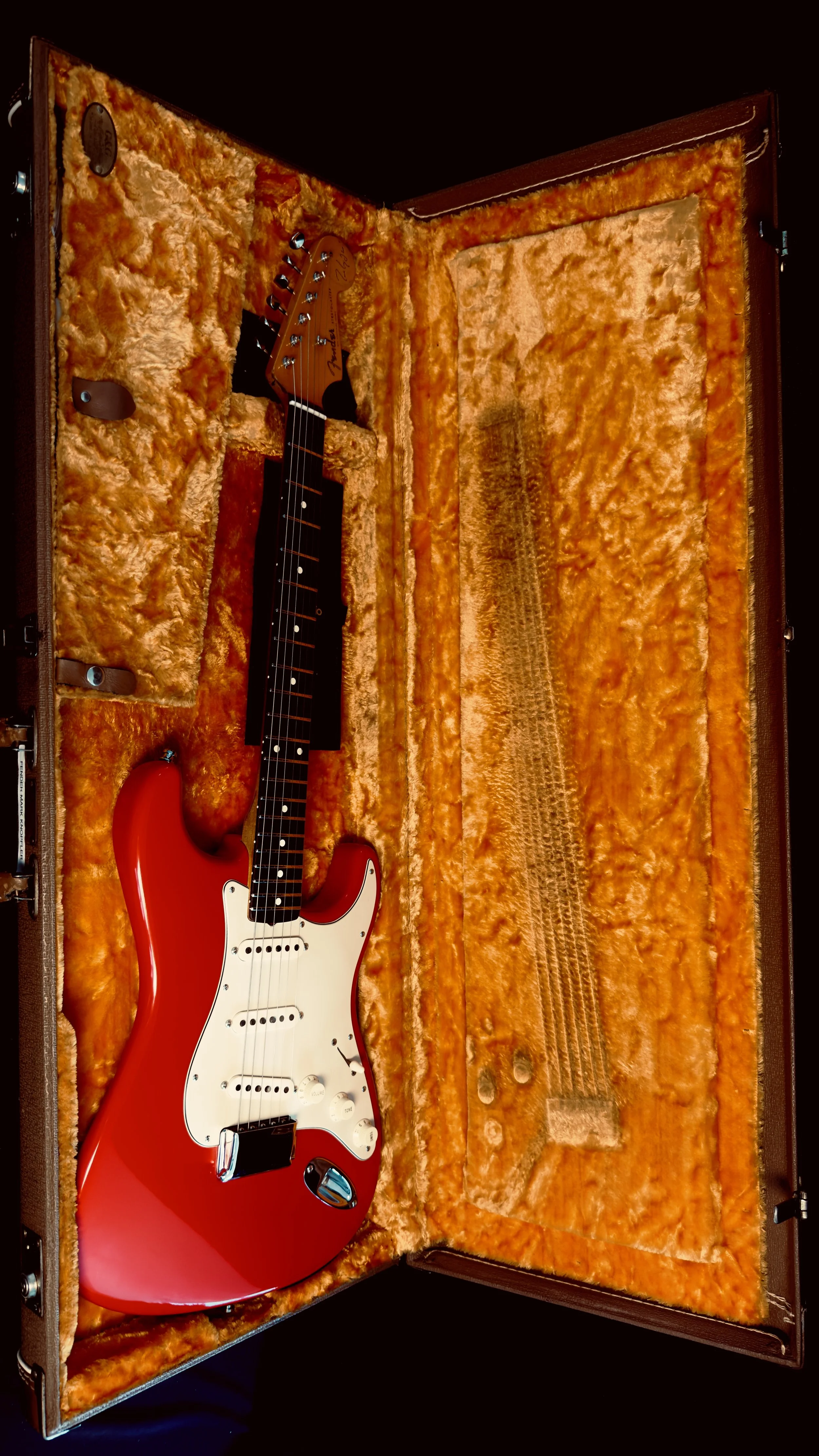 A red electric guitar with a white pickguard inside an orange plush-lined hard case.