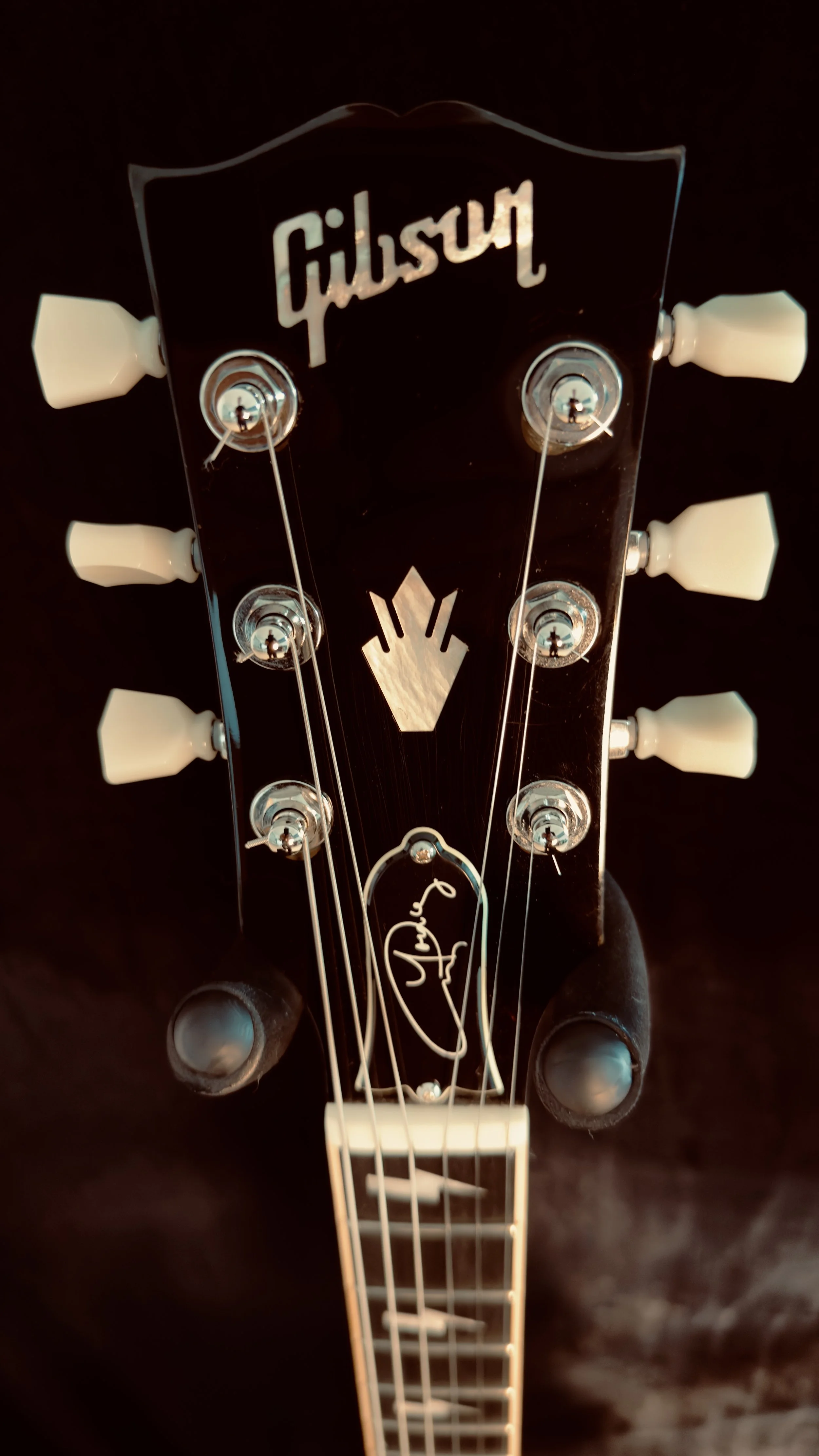 Close-up of a Gibson guitar headstock with tuning pegs, guitar strings, and a signature in white on the black headstock.