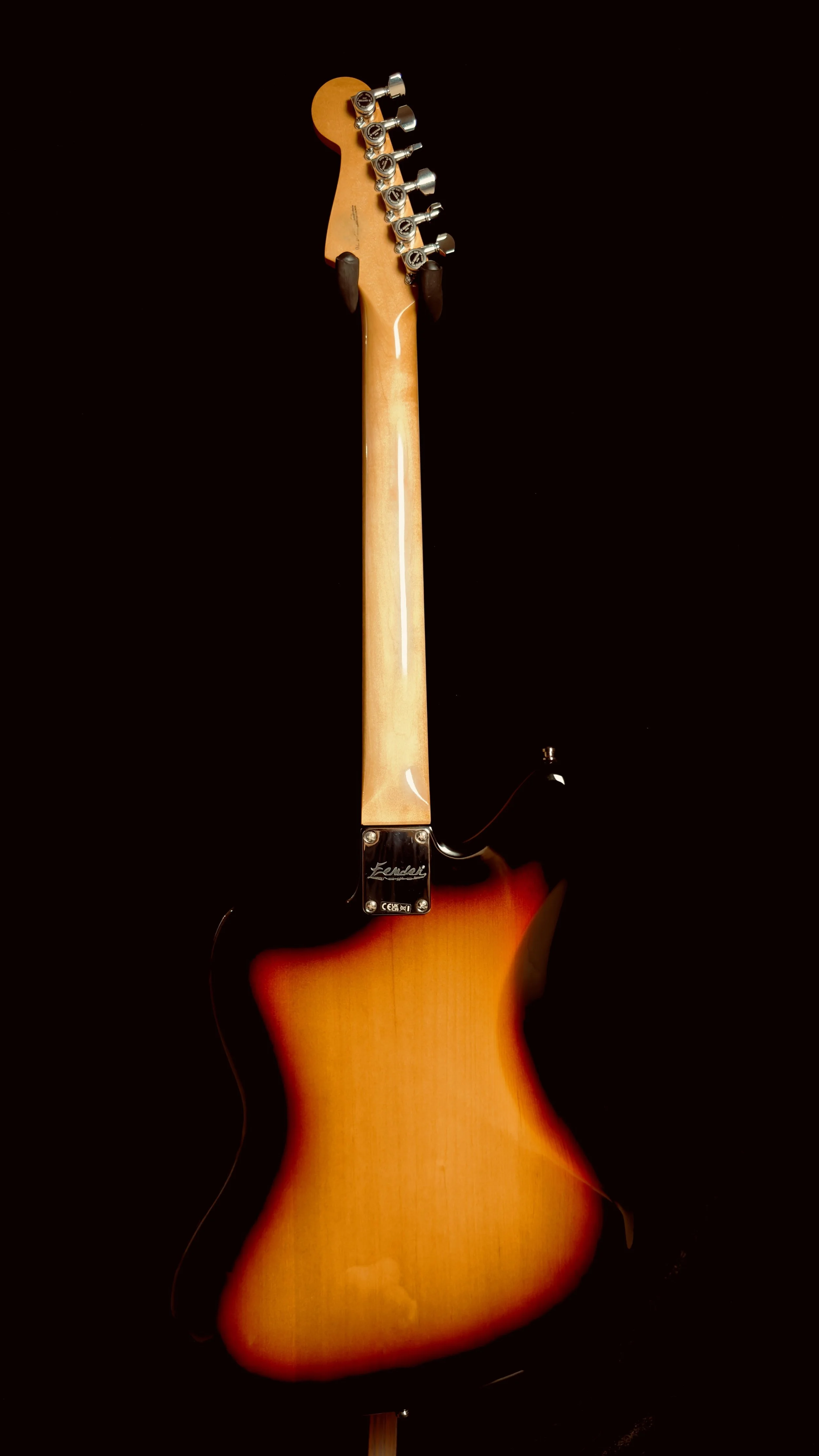 Back of an electric guitar with a sunburst finish, black background, and visible tuning pegs.