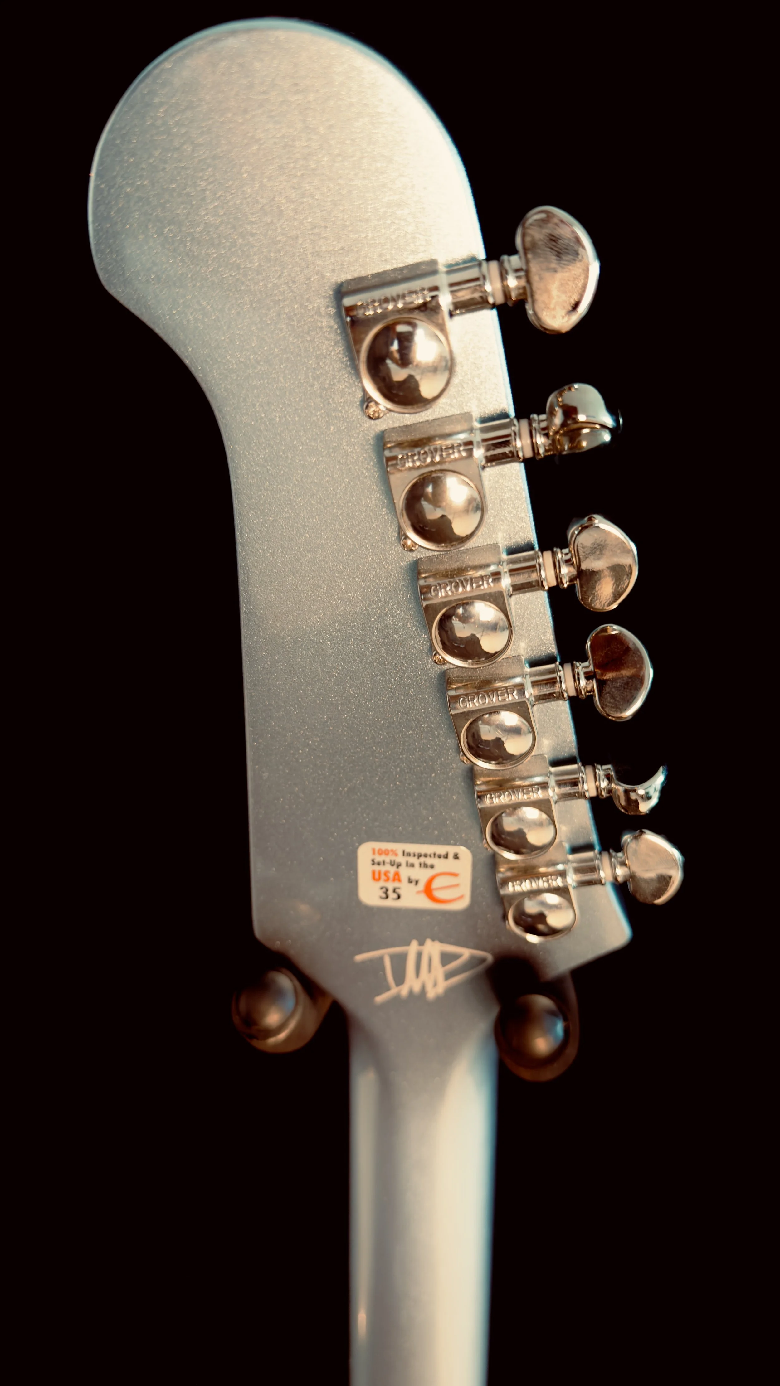 Close-up of the headstock of a silver electric guitar showing six tuning pegs with a sticker that reads '100% inspected & set-up in the USA' and a signature below.