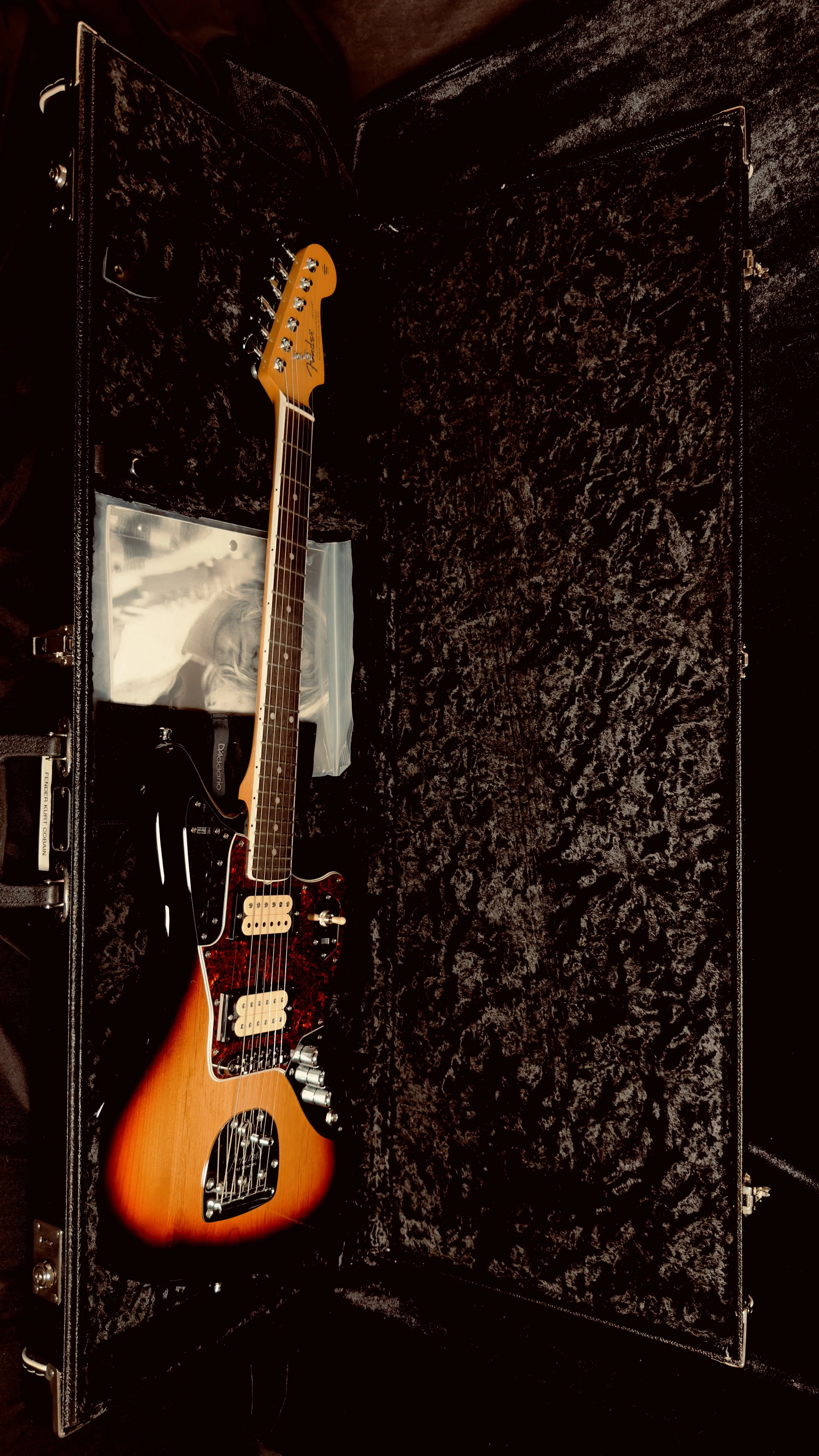 A Fender electric guitar with a sunburst finish and a red and black pickguard inside a black hard case with plush lining.