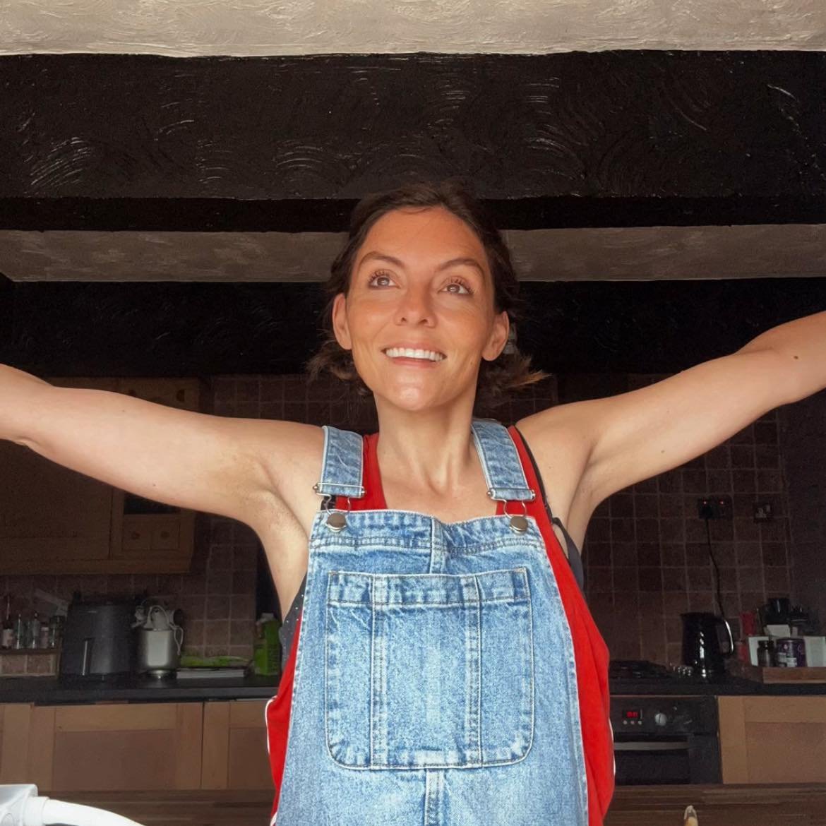 Smiling woman in denim overalls with arms outstretched standing in a kitchen.