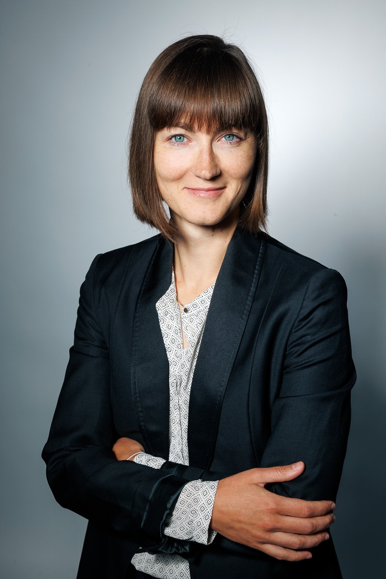 Femme avec cheveux bruns et yeux bleus, portant un blazer noir et une chemise blanche à motifs, souriante, avec bras croisés, contre un fond gris clair.