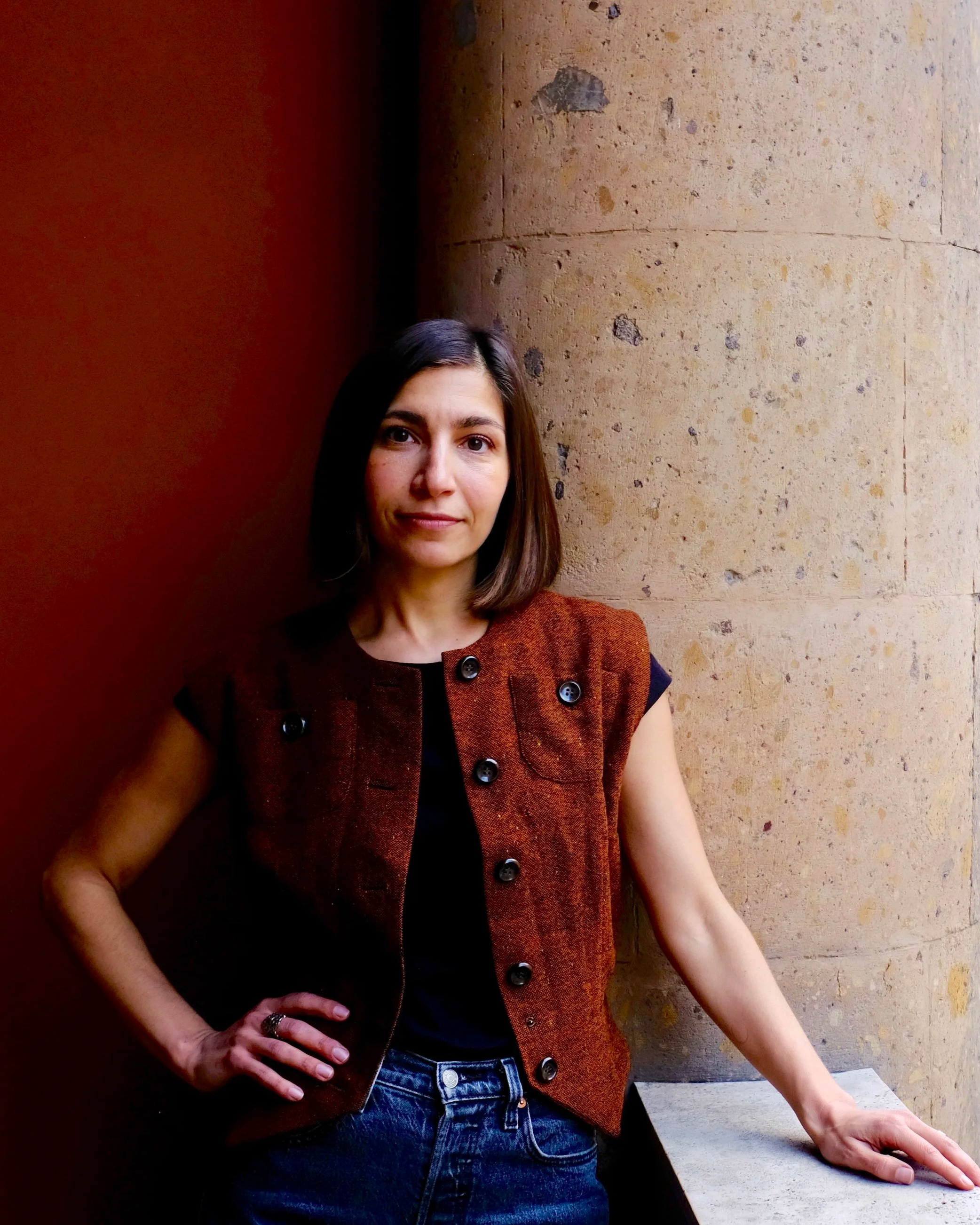 A woman with shoulder-length brown hair, wearing a sleeveless reddish-brown vest over a black shirt, standing with her right hand on her hip and her left hand resting on a ledge, leaning against a stone wall with a textured beige surface.
