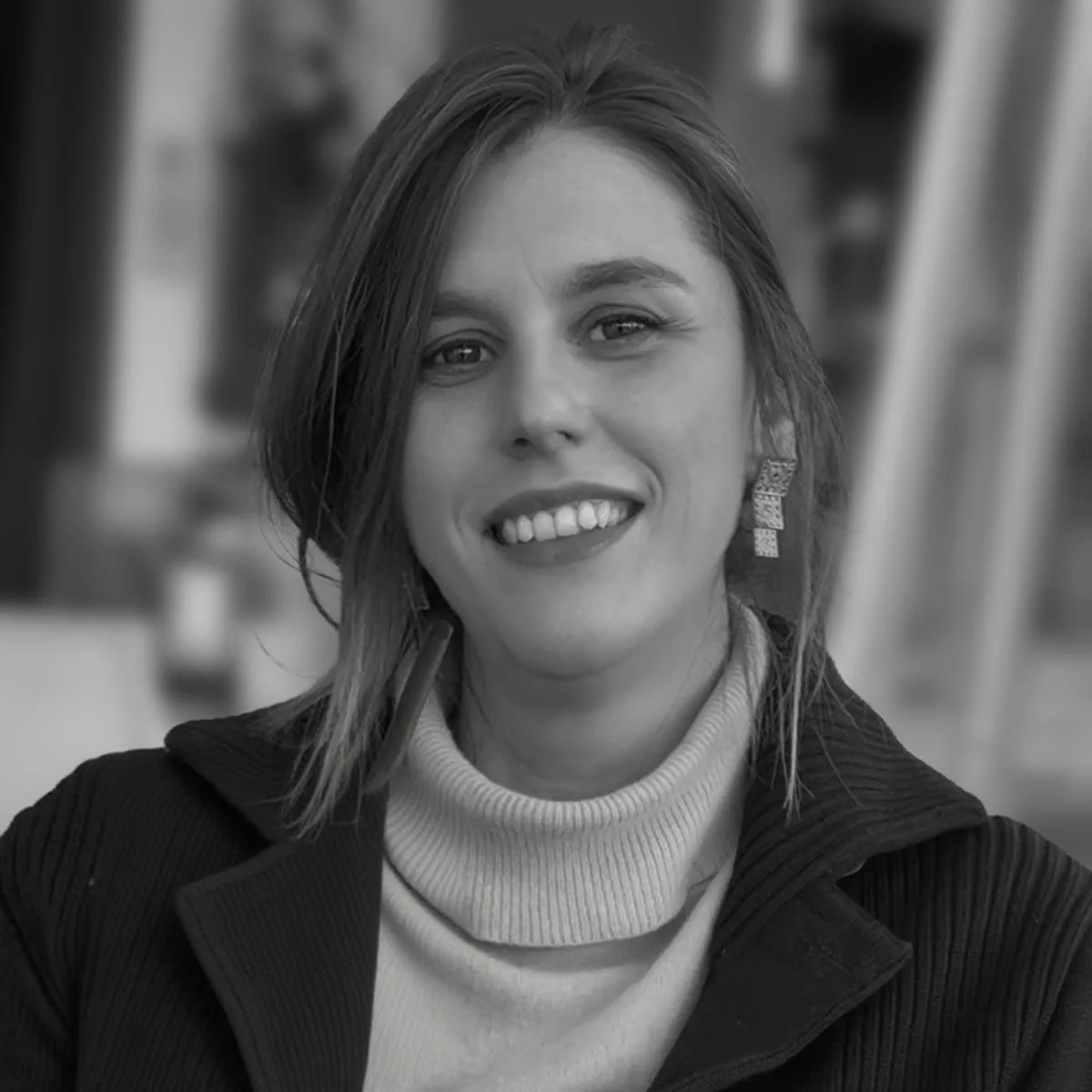 Ragazza che sorride, con orecchini pendenti, con capelli mossi, indossa una maglietta a collo alto e una giacca, sfondo sfocato in bianco e nero.