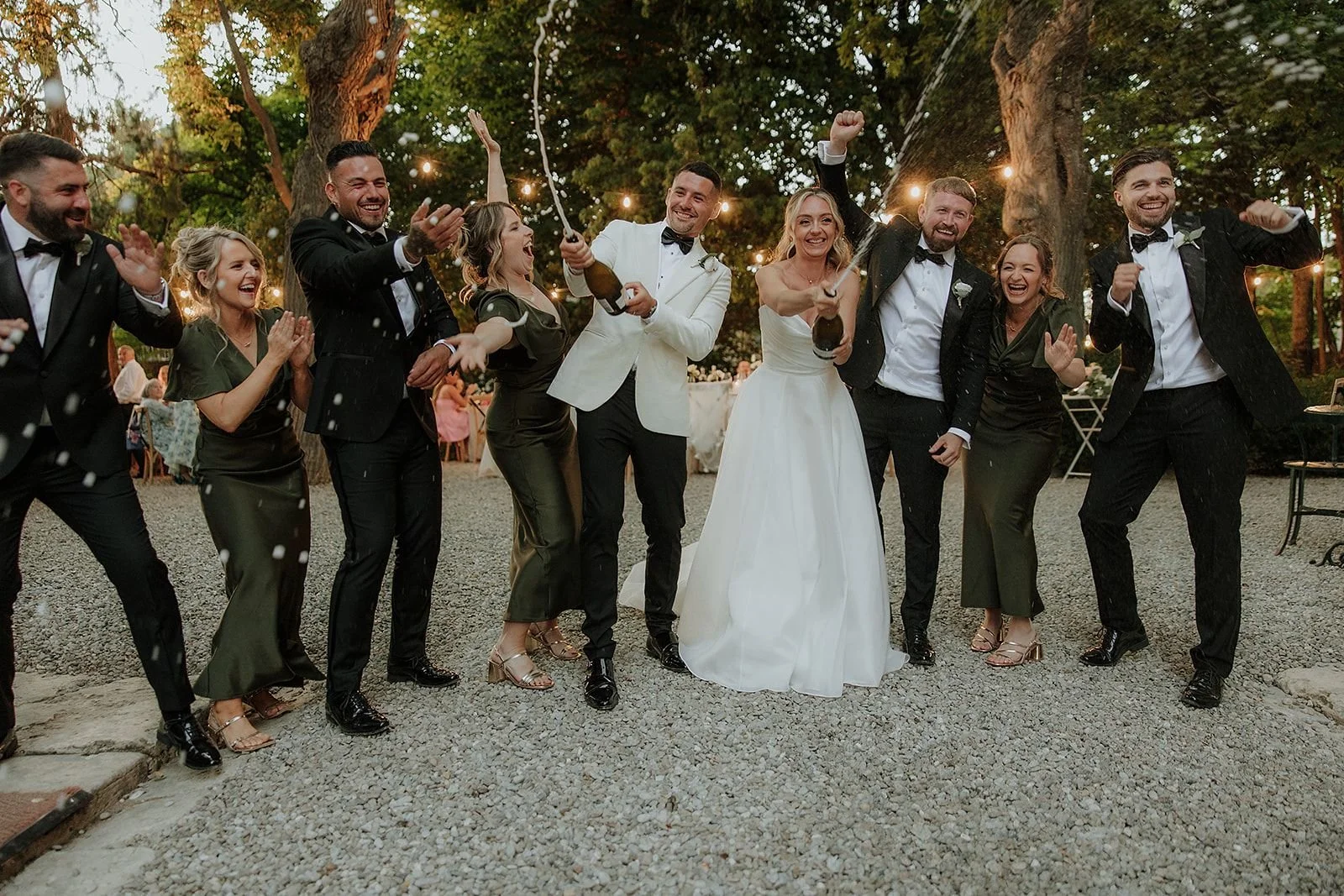 A group of wedding guests celebrating with the bride and groom under string lights in an outdoor setting. The bride is wearing a white wedding gown, and the groom is in a tuxedo. They are joyful, holding bottles, with confetti in the air.