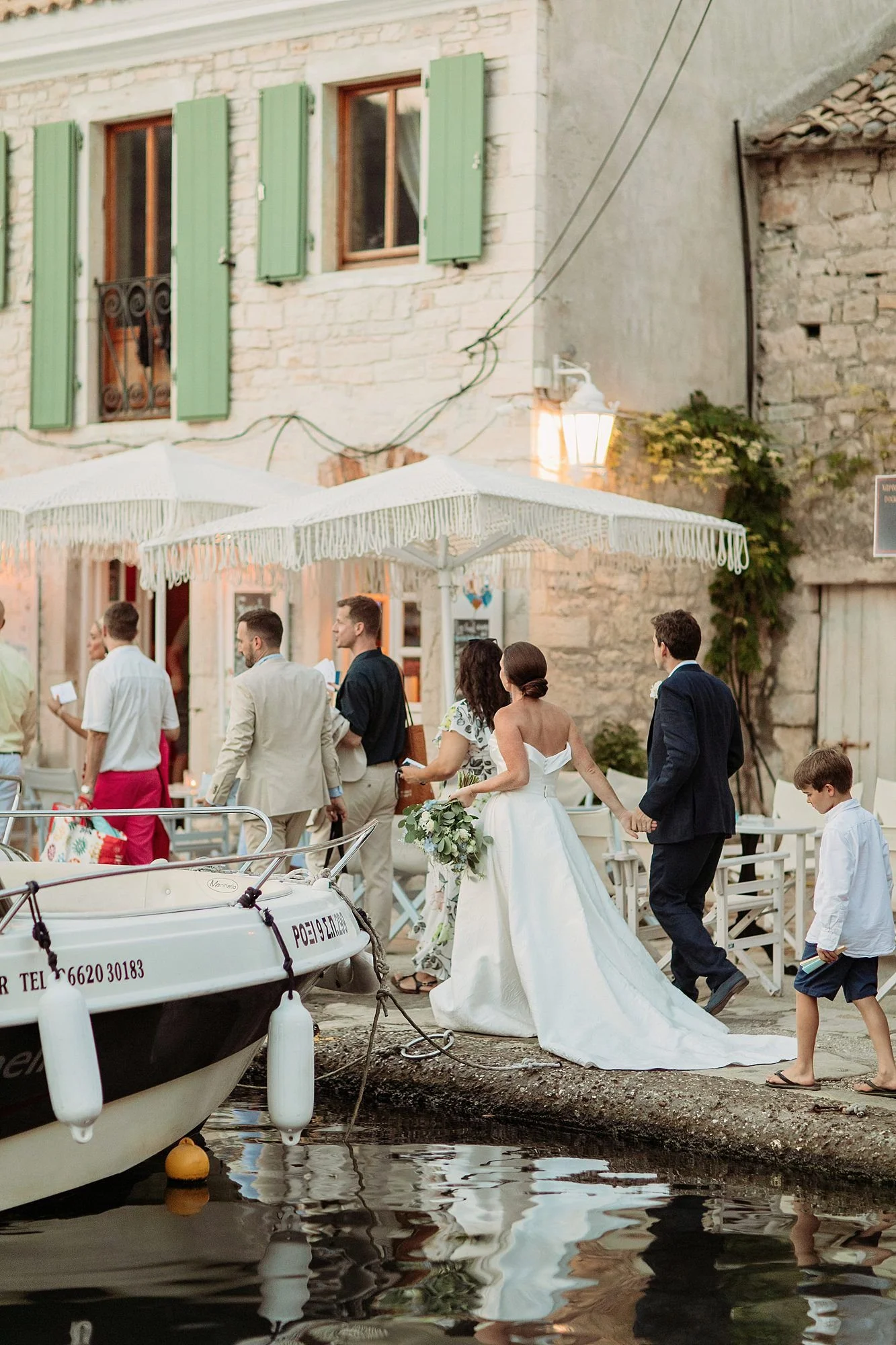 Couple dressed in wedding attire holding hands walking along a waterfront with boats and an outdoor seating area, surrounded by others and rustic buildings with green shutters.