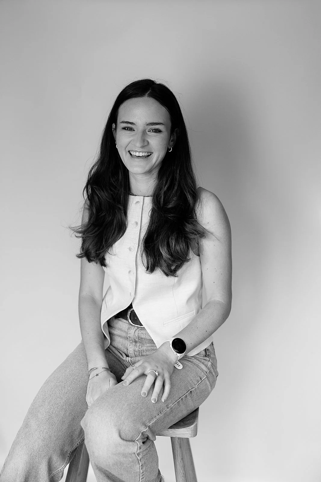 Black and white photo of a young woman with long dark hair, smiling, sitting on a stool against a plain background, wearing a sleeveless top and jeans.