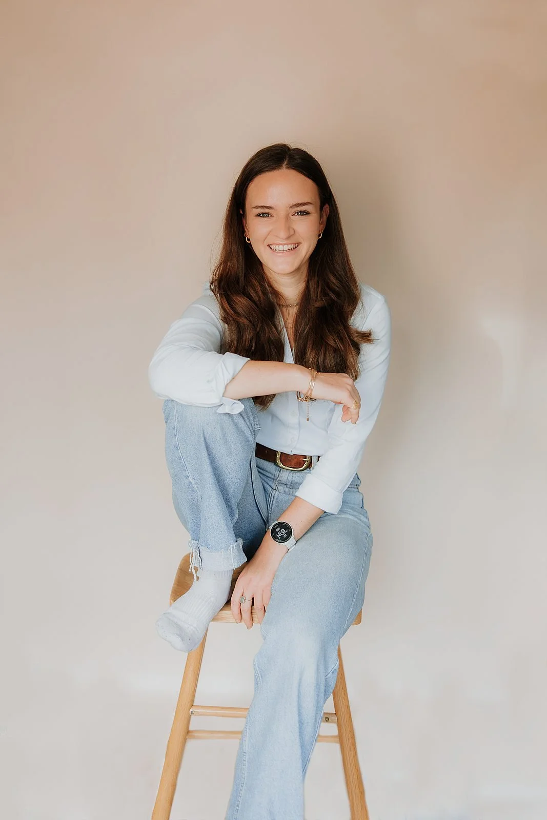 A young woman with long brown hair sitting on a wooden stool against a plain beige background, smiling at the camera. She is wearing a white button-up shirt, light blue ripped jeans, white socks, and a smartwatch.