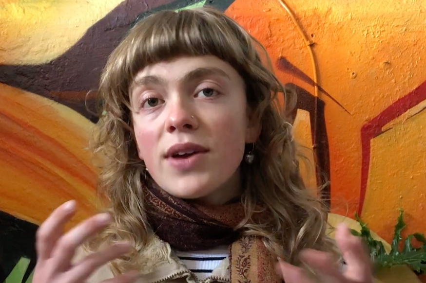 A woman with wavy light brown hair, light skin, wearing a nose ring and earrings, and a brown scarf around her neck, speaking or explaining something inside a room with orange and yellow painted walls.
