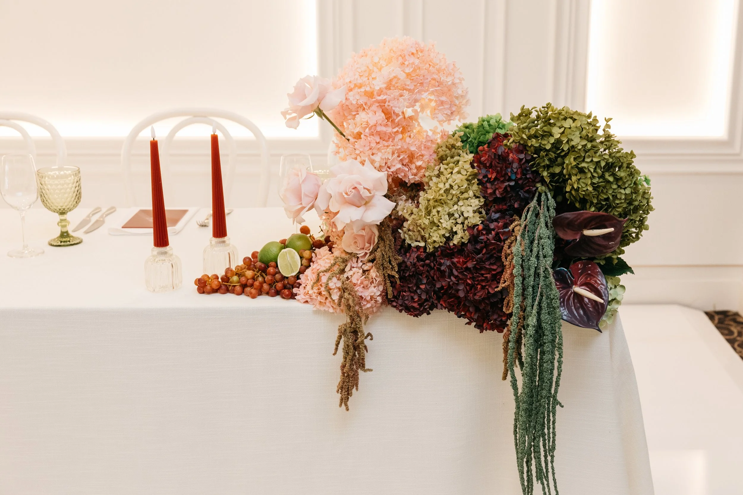 Decorative floral centerpiece with pink and green hydrangeas, roses, calla lilies, grapes, and limes on a table with red candles in glass holders, set for a formal event.