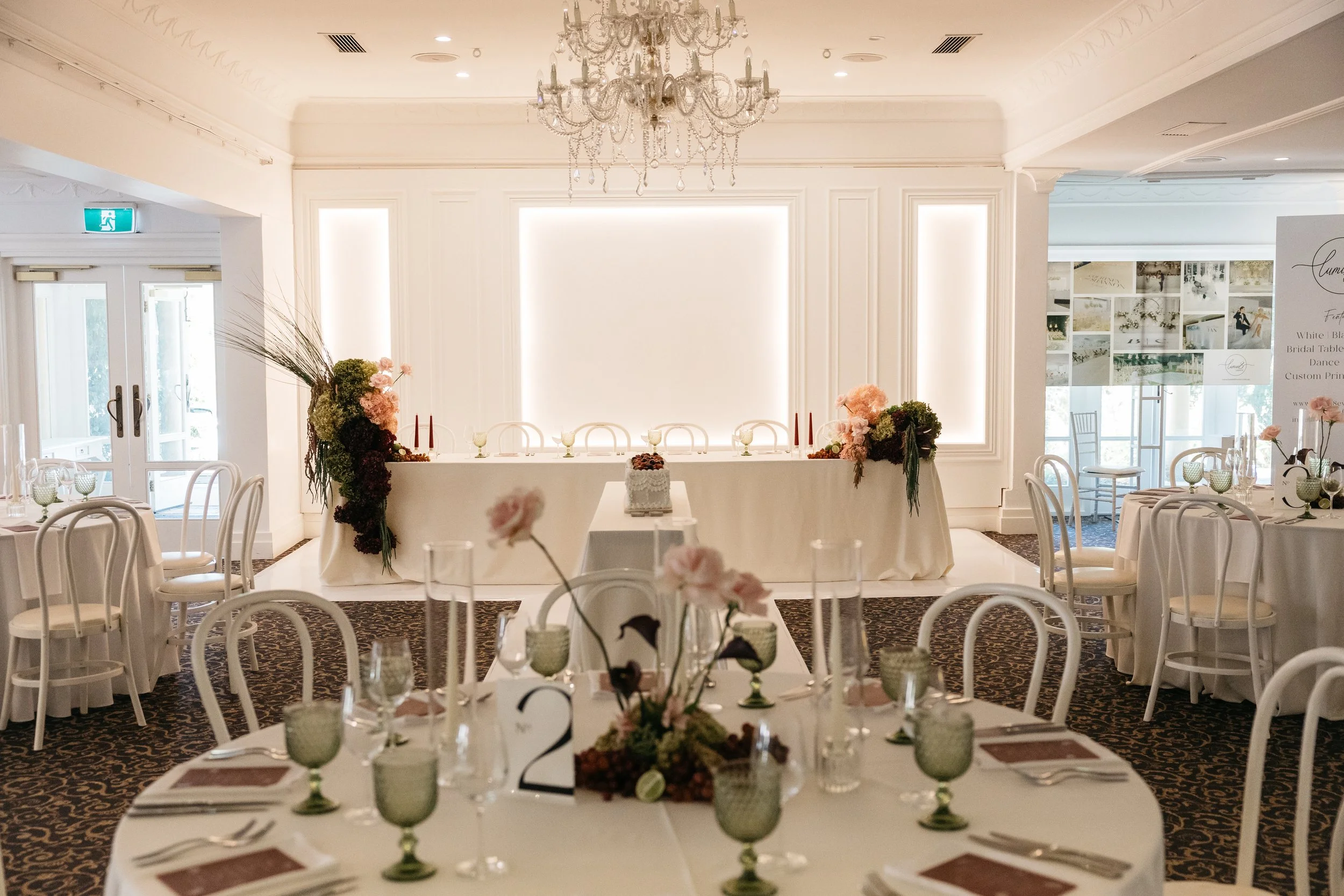 Elegant banquet hall decorated for a wedding reception with tables set, floral centerpieces, and a head table with candles and flowers, illuminated by soft lighting and a chandelier.
