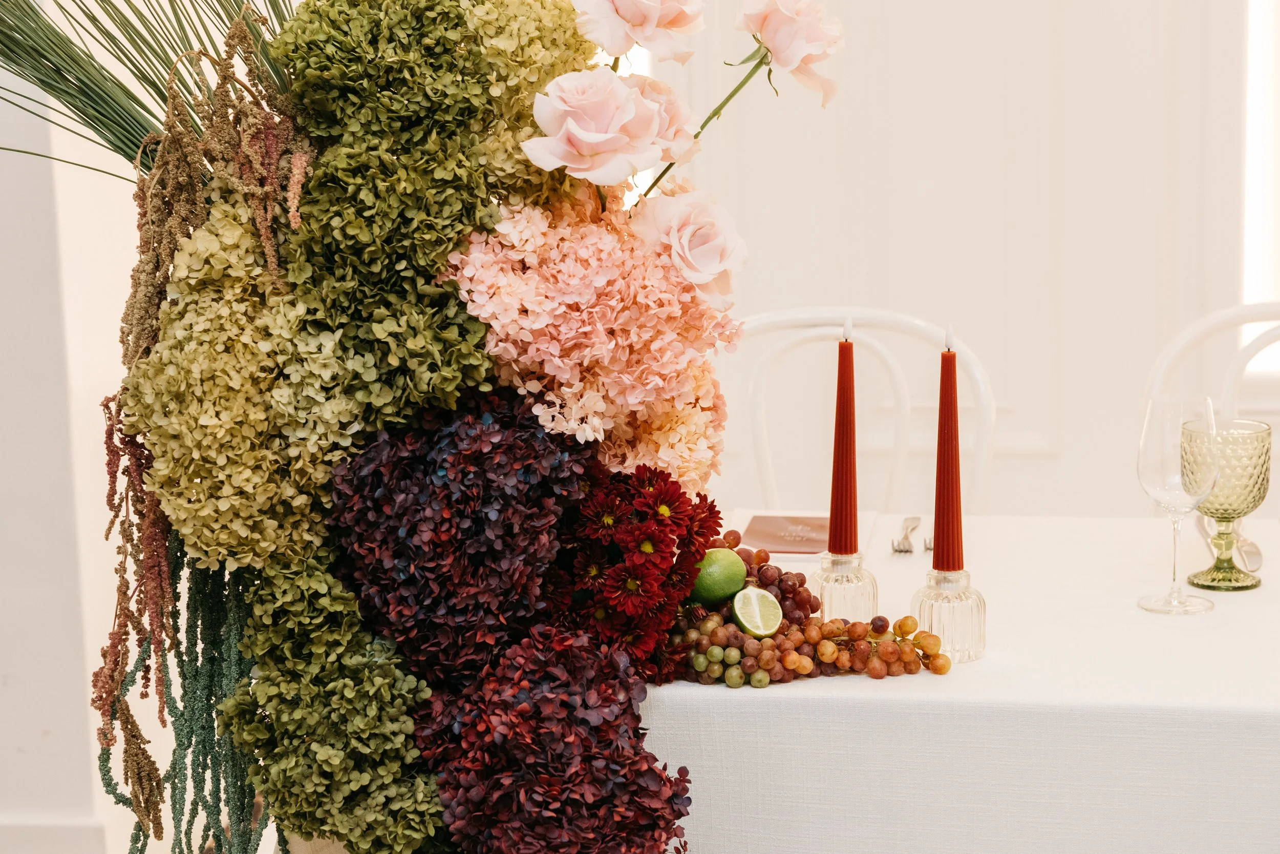 A floral arrangement featuring various hydrangeas and roses, arranged on a table with red taper candles, citrus fruits, and glassware in a bright, elegant setting.