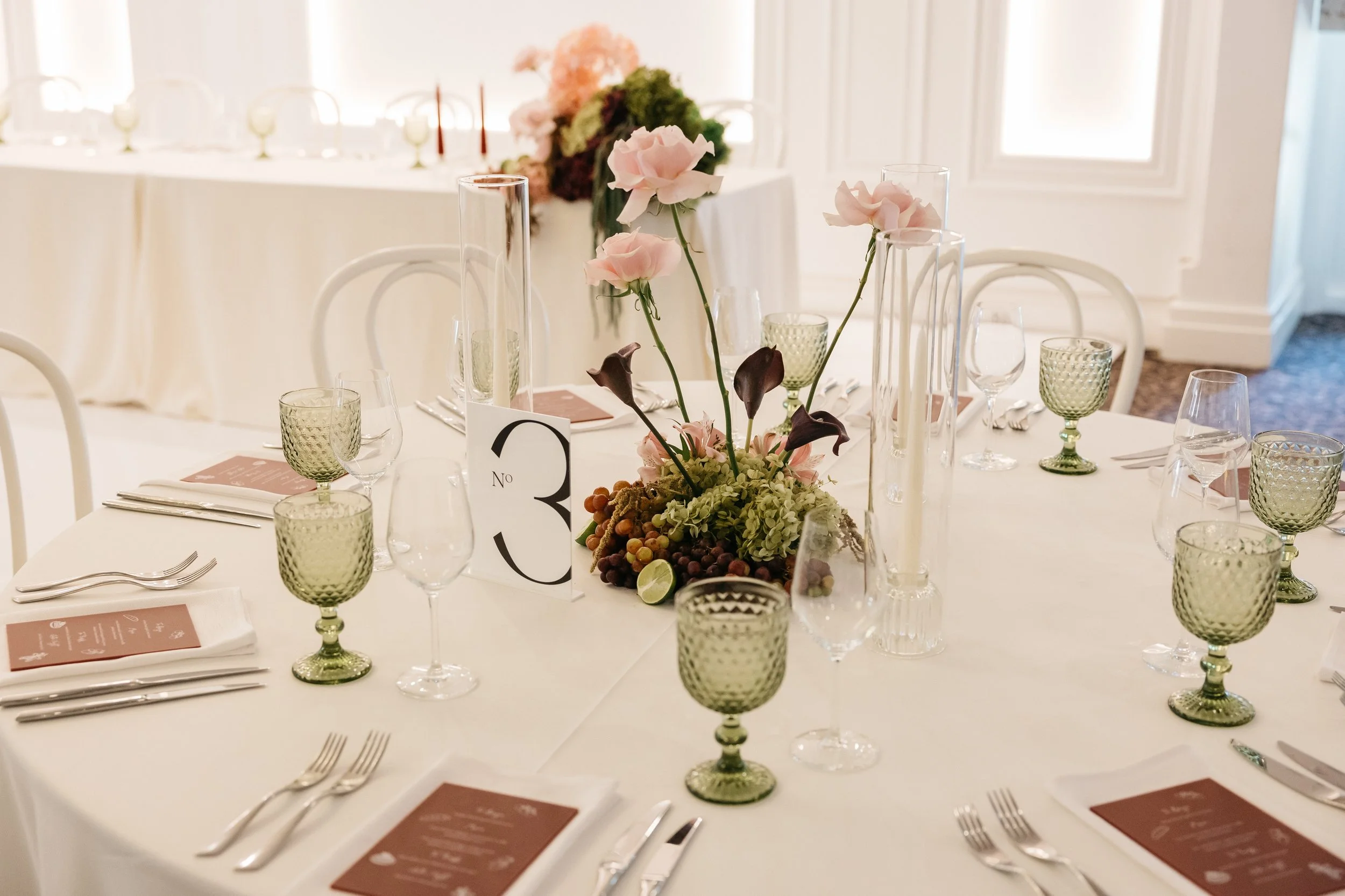Elegant banquet table set with floral centerpiece, green glasses, wine glasses, and place settings in a bright, white room.