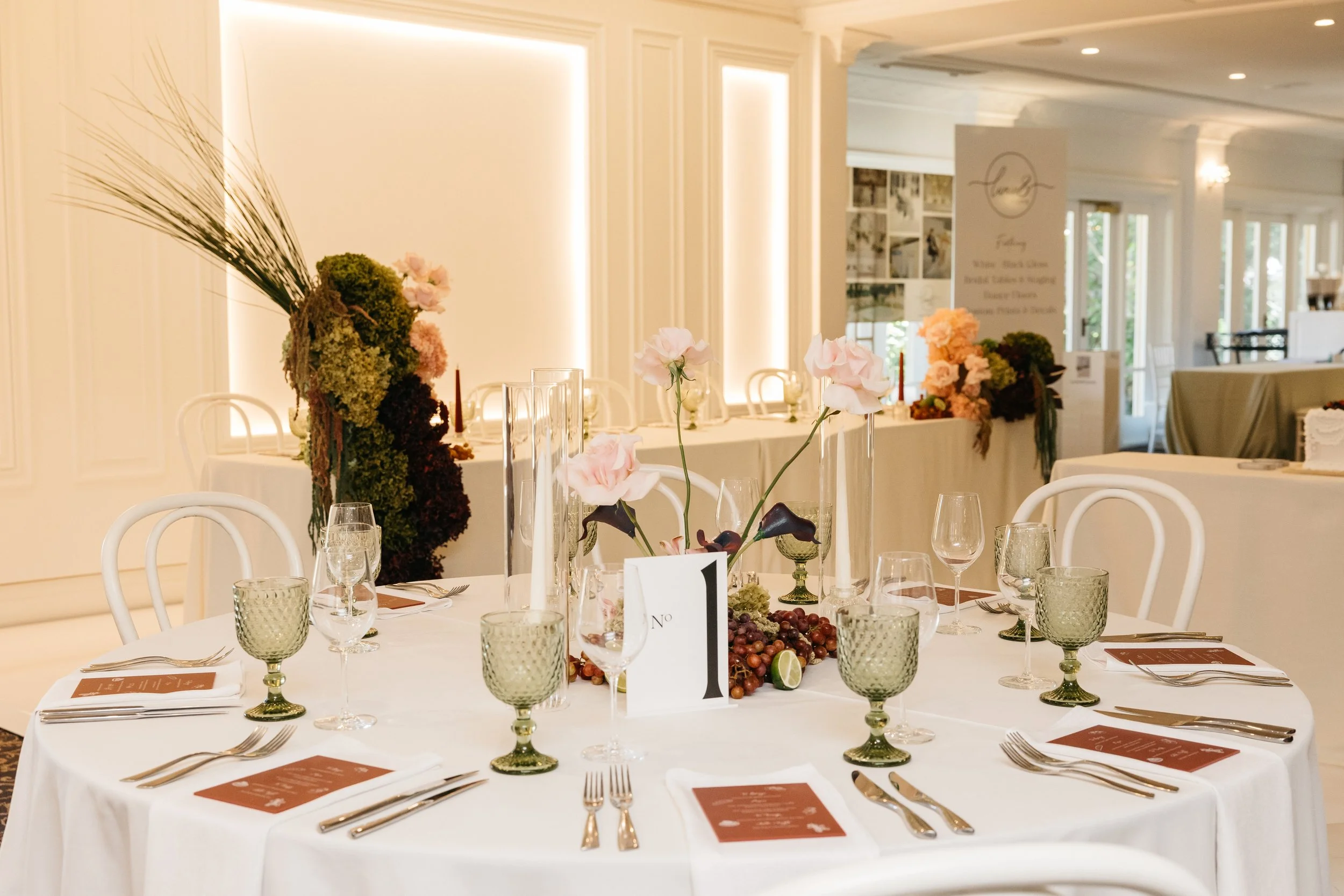 Elegant wedding reception table decorated with pink flowers, green glassware, and a table number card in a bright, well-lit room with white walls and chairs.