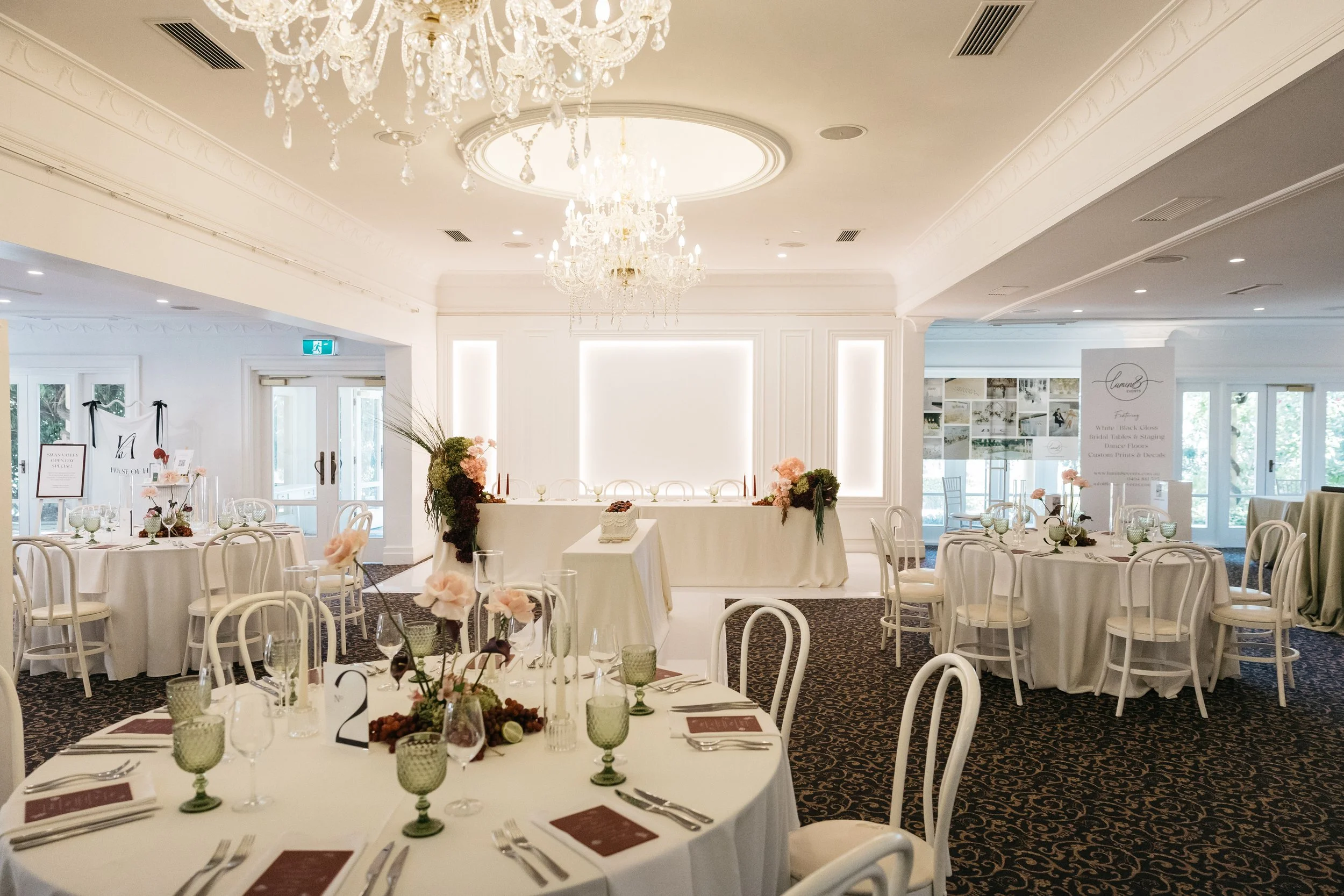 A decorated banquet hall with white walls, draped tables with white tablecloths, floral centerpieces, and elegant chairs, set up for a formal event or wedding reception.