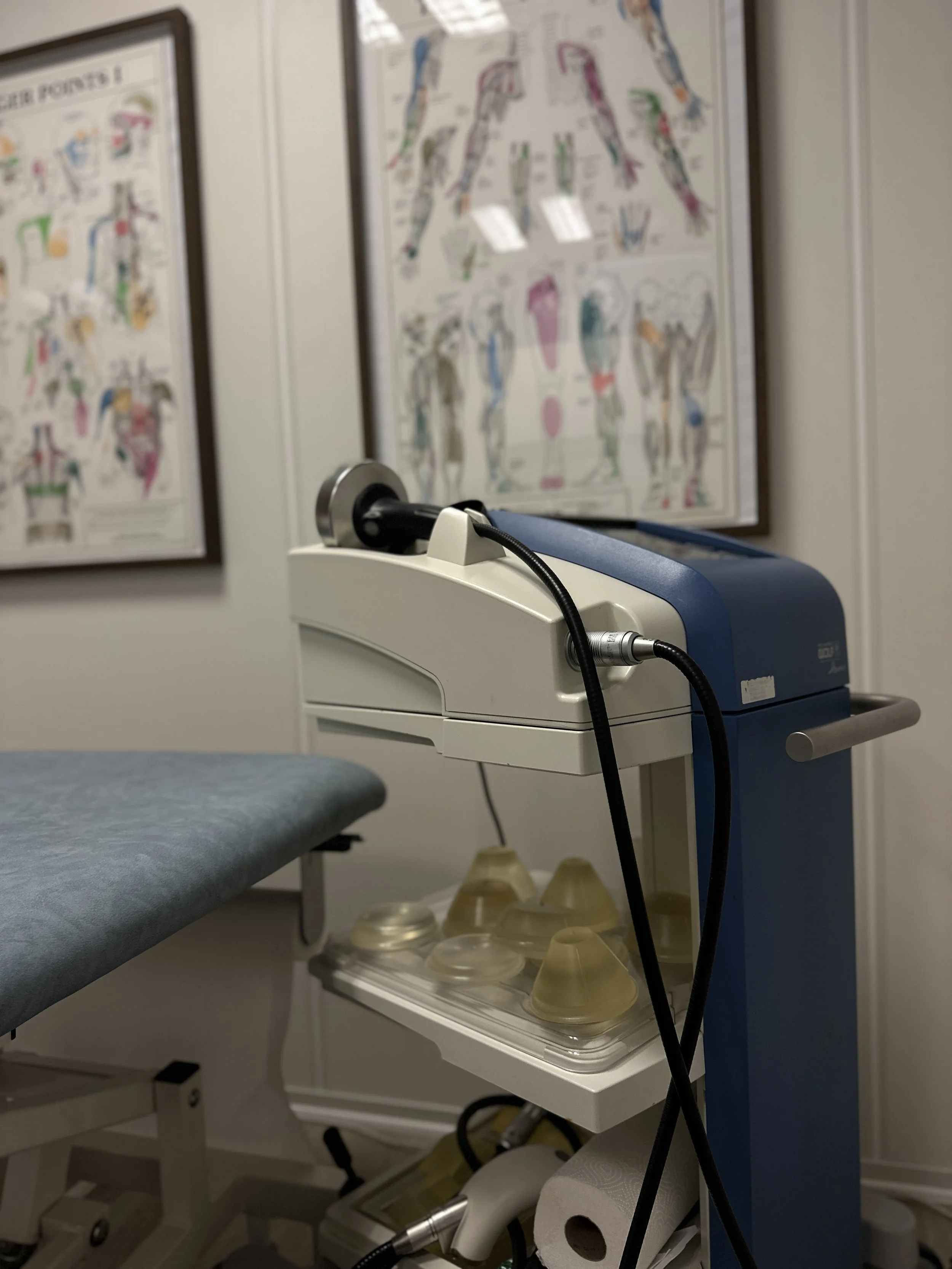 Medical ultrasound machine next to examination table in a clinic room with anatomical charts on the wall.
