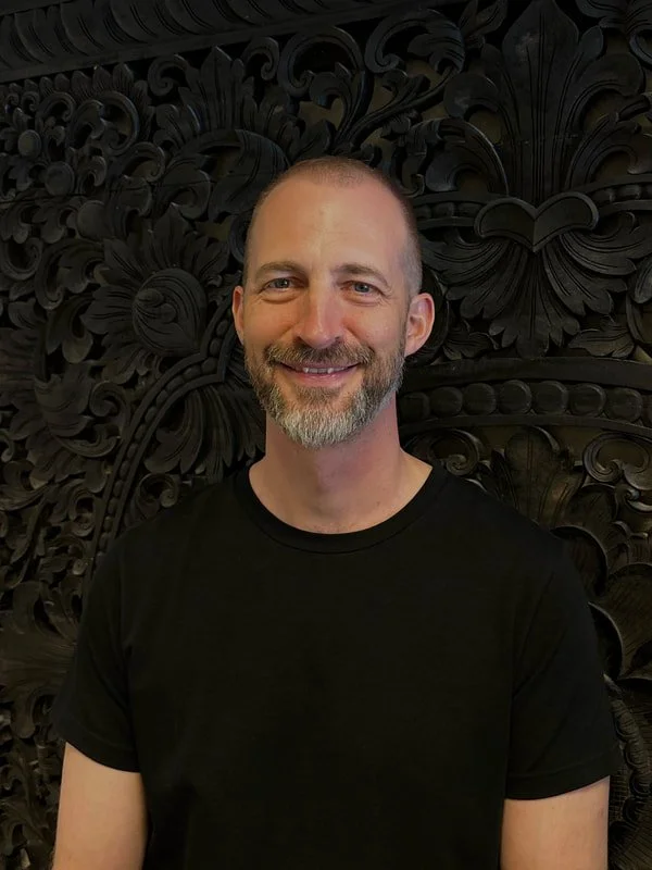 A man with a beard smiling, wearing a black t-shirt, standing in front of an intricately carved dark wooden wall.