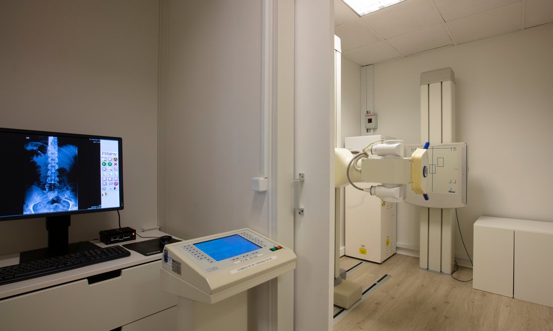 Medical imaging room with X-ray machine, monitor displaying a human pelvis X-ray, and computer equipment.