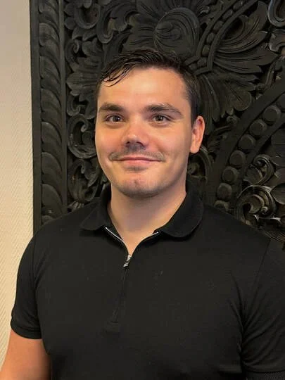 A young man with dark hair and fair skin wearing a black polo shirt, standing in front of an ornate, dark wooden carved background.