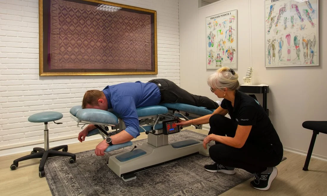 A chiropractor performs an adjustment on a patient lying face down on a specialized chiropractic table in a clinic. The chiropractor is kneeling beside the table, operating a control panel. The room has medical diagrams on the wall and a side table with model of a spine.