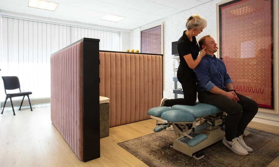 A chiropractor adjusting a man's neck in a treatment room with a privacy wall and chairs.