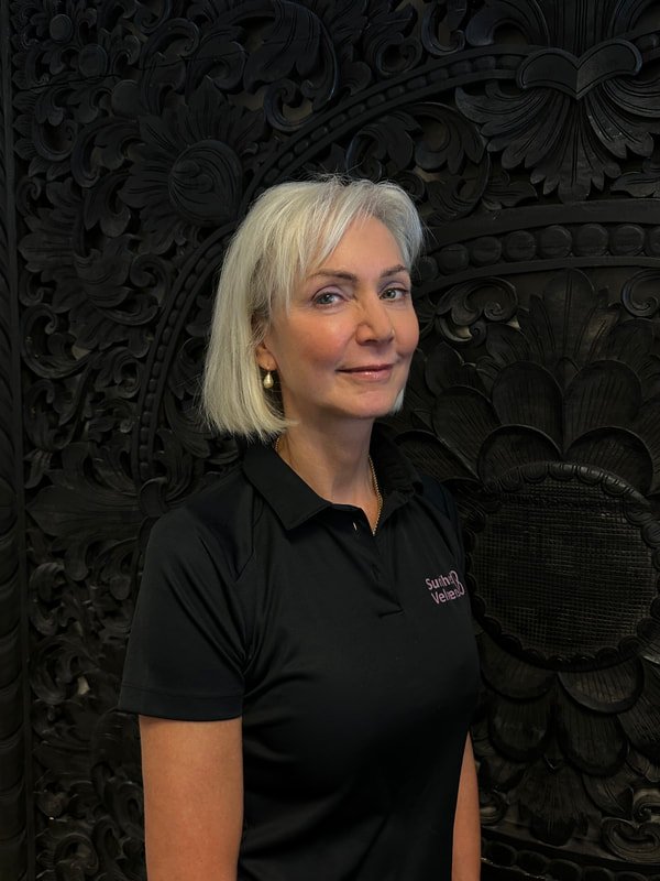 A woman with shoulder-length white hair and earrings wearing a black collared shirt with a logo, standing in front of a dark ornate carved background.