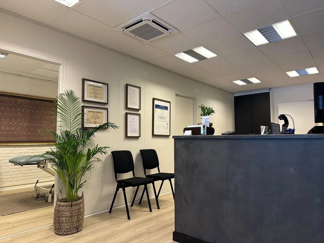 Waiting room or reception area with two black chairs, a potted plant, framed certificates on the wall, a reception desk with a small plant, and a window with a curtain.