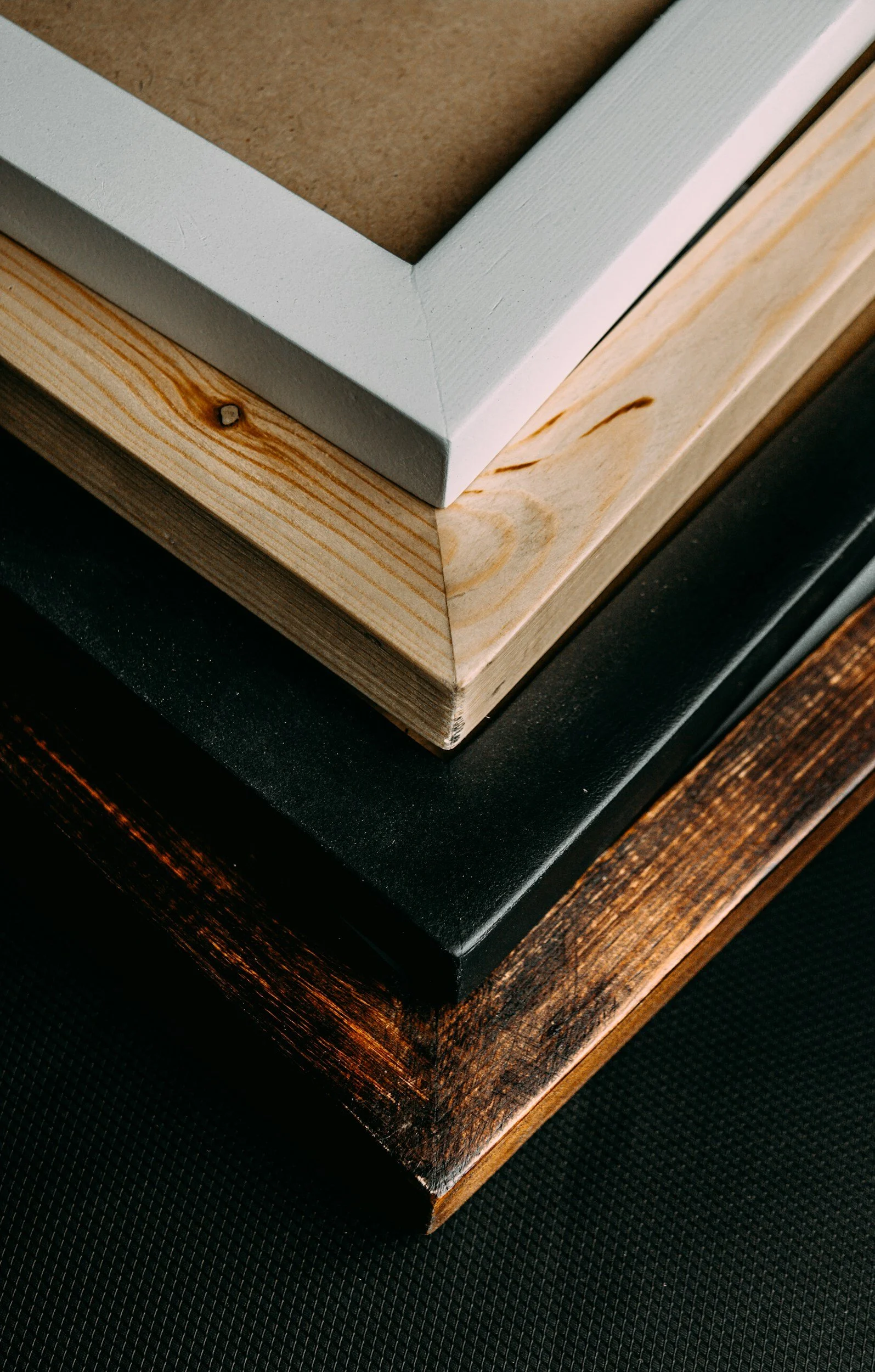 Close-up of three picture frames with different colored borders — white, natural wood, and black — resting on a dark textured surface. The frames are stacked with the smallest on top and the largest at the bottom.