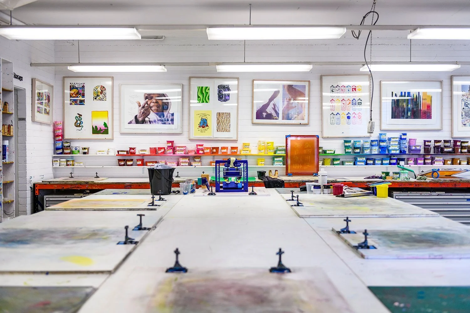 An art or printmaking studio with large tables, colorful paint supplies on shelves, and framed artwork on the white brick walls.