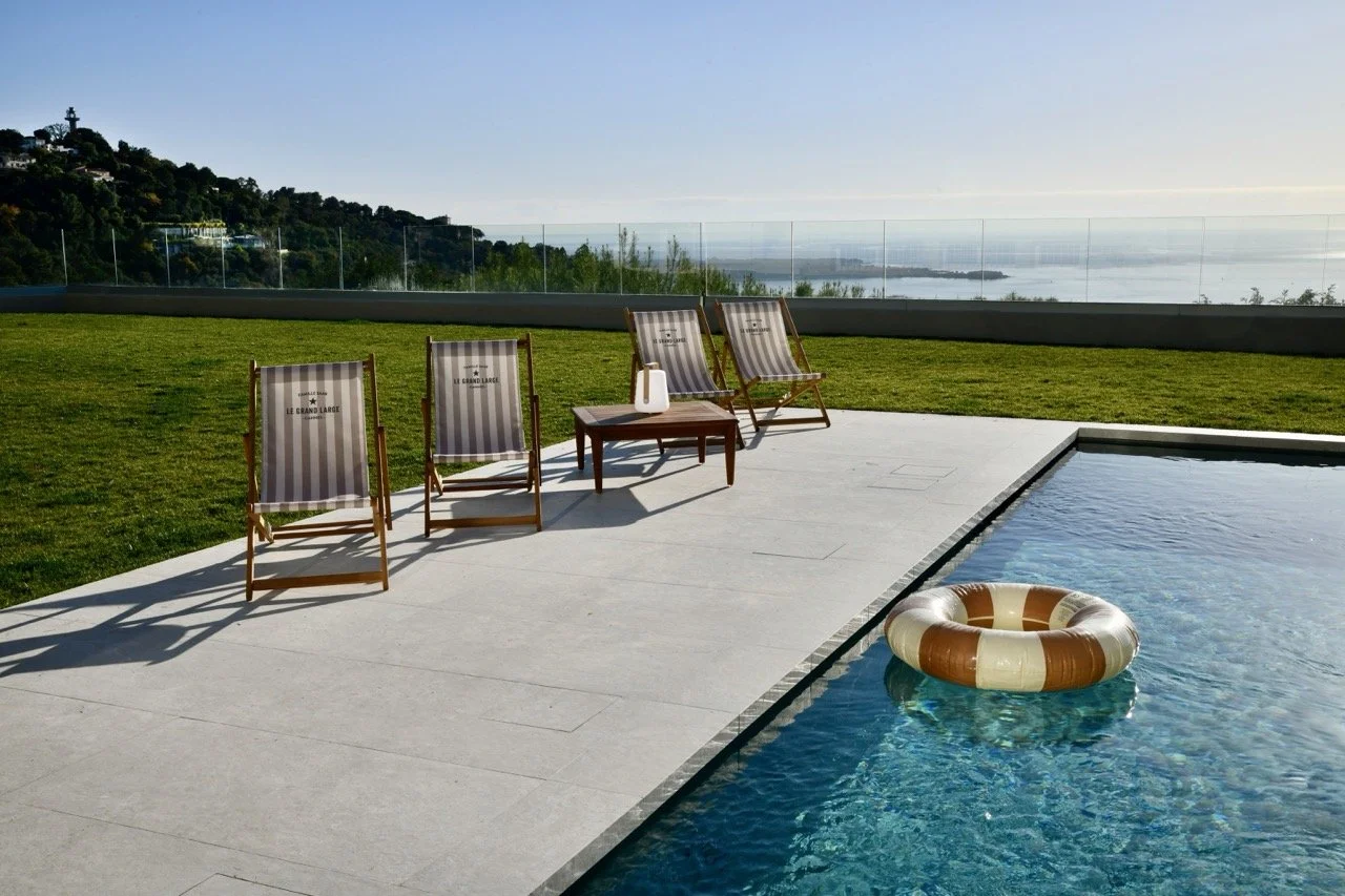 Chaises longues au bord d'une piscine avec un paysage marin en arrière-plan, un porte-gobelet et une bouée dans l'eau.