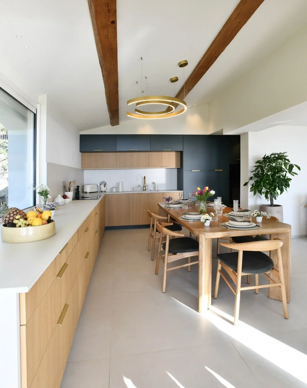 Cuisine moderne avec plan de travail en blanc, armoires en bois clair et en bleu foncé, table à manger en bois avec six chaises, décoration florale, fruits dans un bol, grande fenetre laissont entrer la lumière.