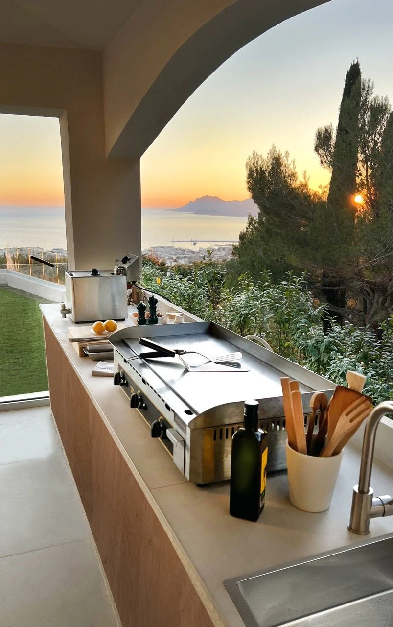 Cuisine en plein air avec plancha électrique, bouteille d'huile d'olive, ustensiles en bois, lampe à citron, quelques citrons et un magnifique coucher de soleil sur la mer et les montagnes en arrière-plan.