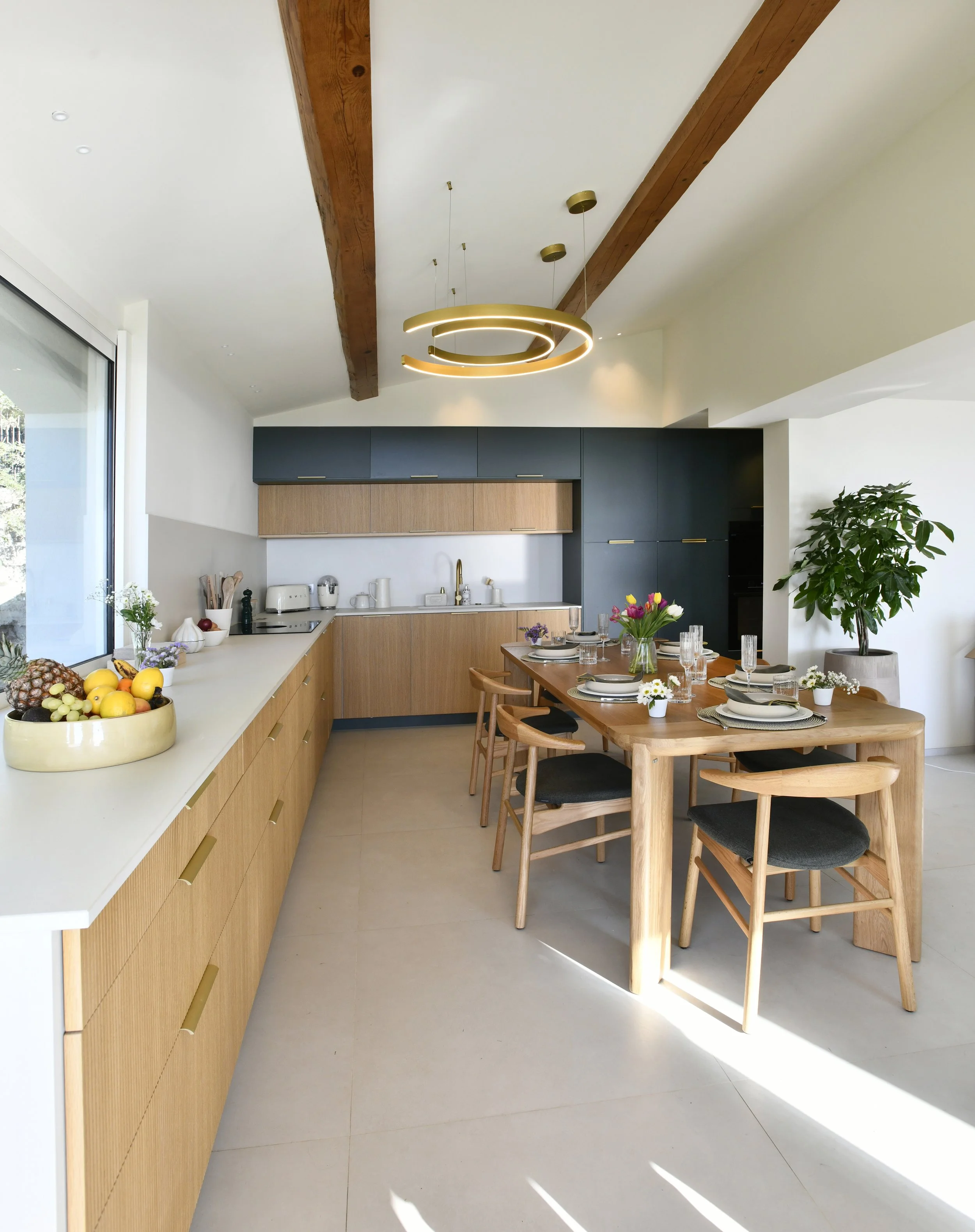 Cuisine moderne avec plan de travail en blanc, armoires en bois naturel, suspendu bleu marine, table à manger en bois avec six chaises en bois avec assises noires, une grande plante verte, éclairage suspendu rond moderne, et une grande fenetre laissa