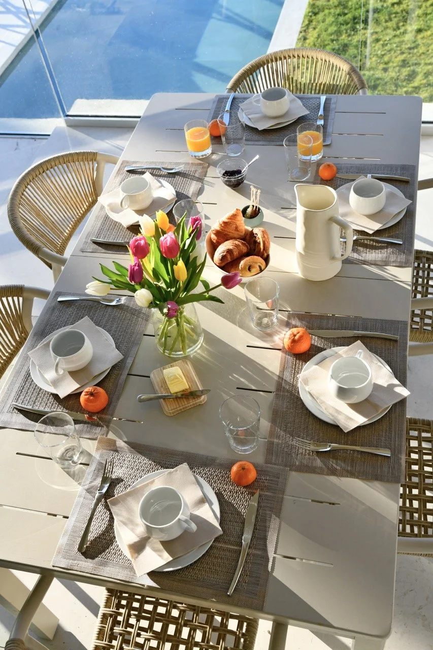 Table de petit-déjeuner avec des assiettes, tasses, verres, couverts, un vase de tulipes, une carafe, des pains, des fruits et une petite boîte de beurre, dans une salle lumineuse avec vue sur l'extérieur.