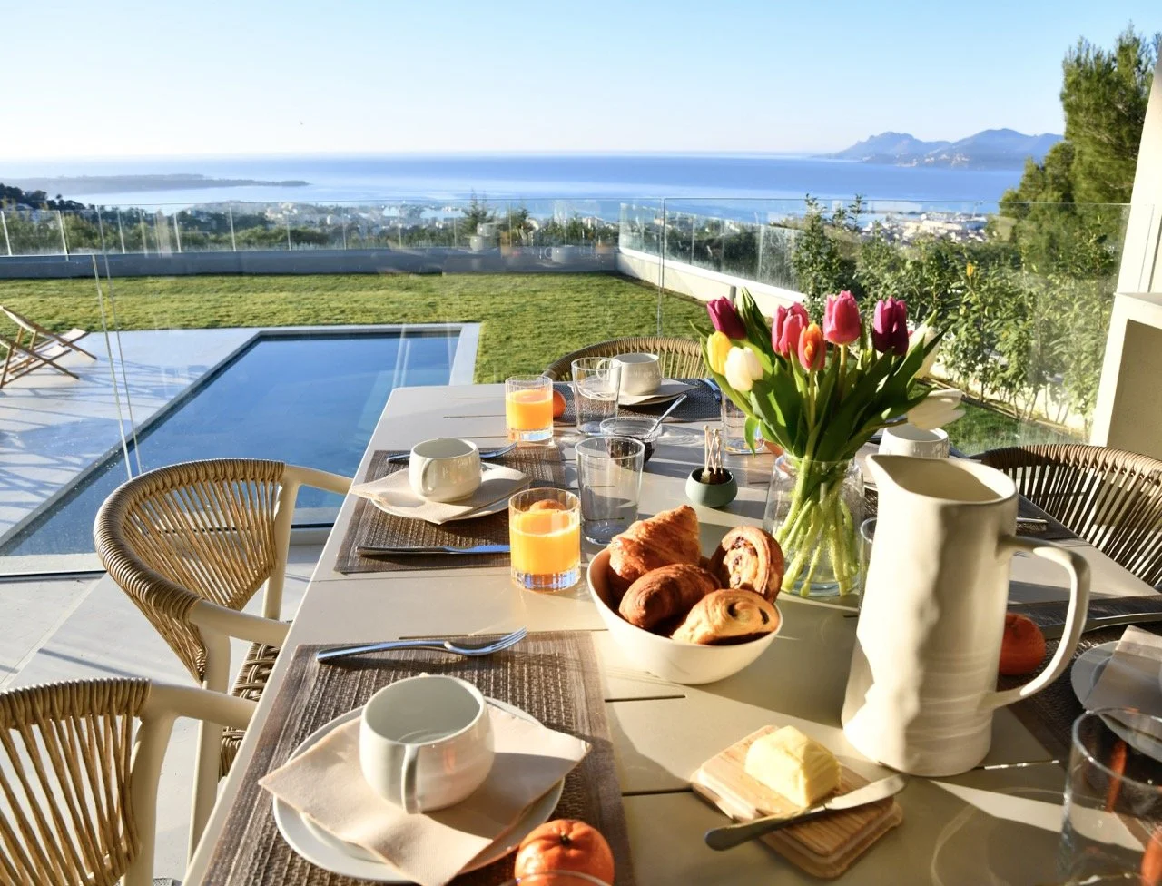 Table de petit-déjeuner avec des croissants, des boissons, une carafe, un vase avec des tulipes, dans une salle avec vue sur la mer et un jardin.
