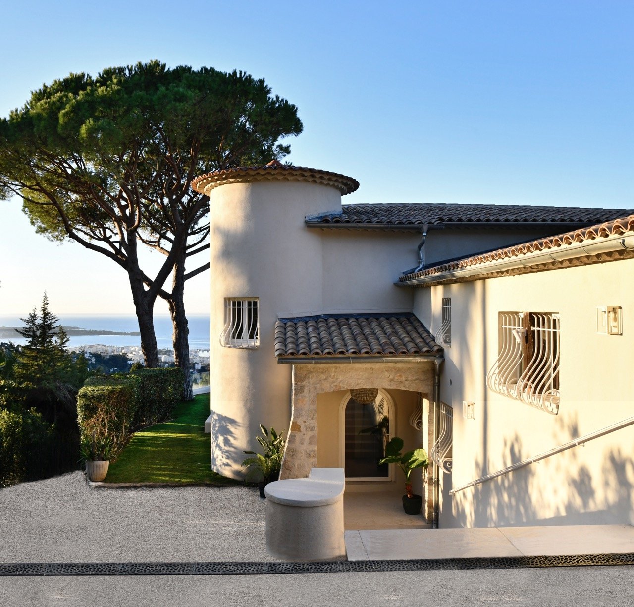 Une maison avec un mur blanc, des fenêtres avec des barreaux, un toit en tuiles, un arbre grand et touffu, une allée en gravier, et un paysage urbain au loin avec de l'eau.